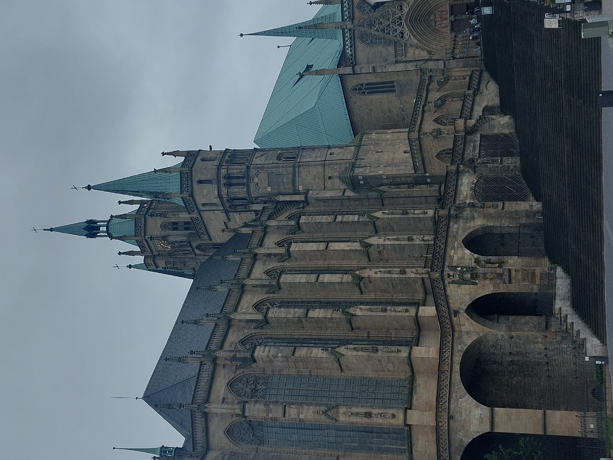 The image shows a large, Gothic-style cathedral with multiple spires, constructed of stone. The cathedral has a long, pitched roof covered in a greenish-blue material. Large, arched windows with intricate tracery are visible along the side of the building. In the foreground, a wide, dark staircase leads up to the entrance of the cathedral. Several people are visible at the top of the stairs, near the entrance. The sky is overcast and gray. This is the Erfurt Cathedral, also known as St. Mary's Cathedral, located in Erfurt, Germany.
