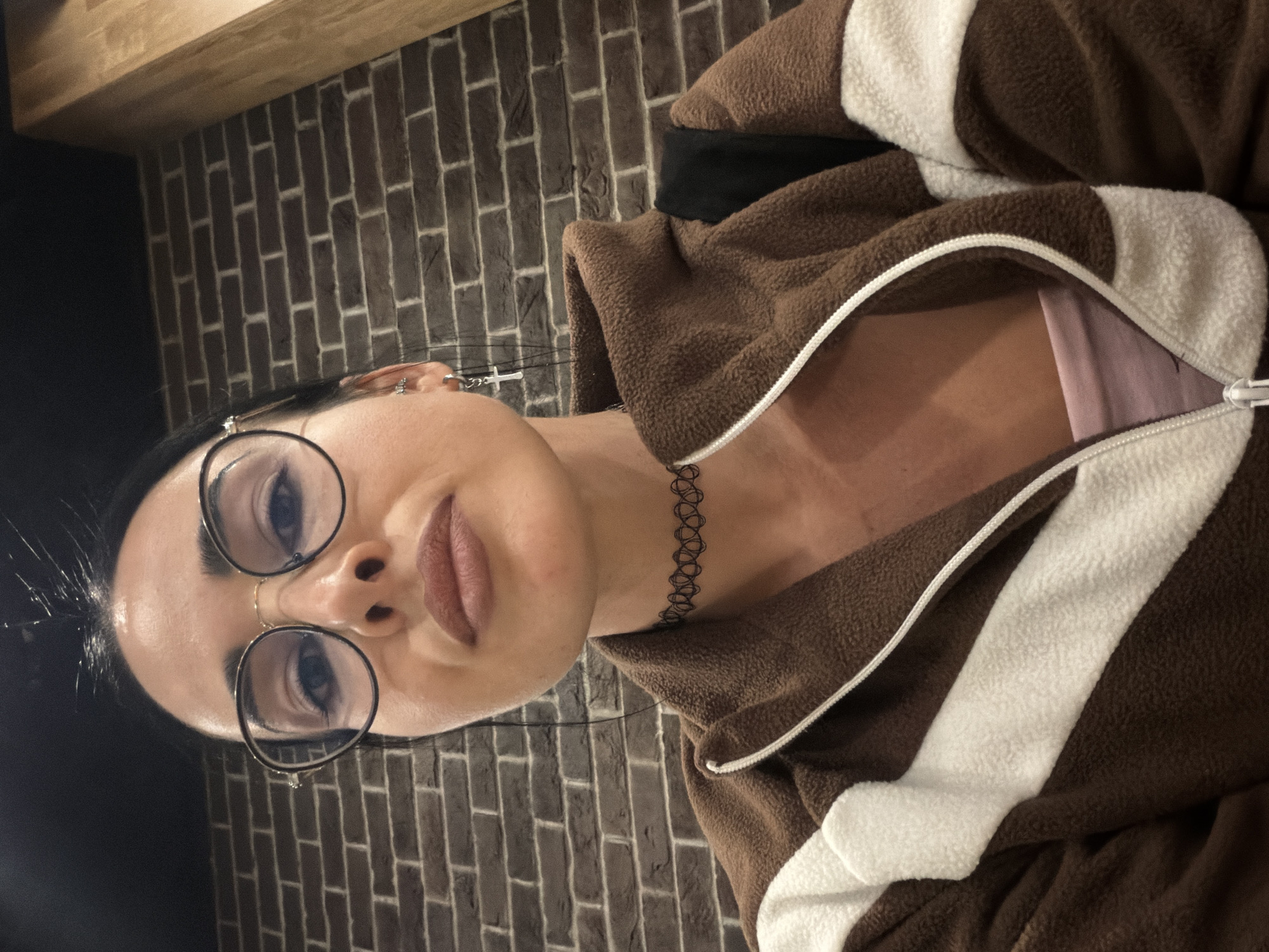 A portrait-style photograph, rotated 90 degrees clockwise, features a person looking directly at the camera. The subject, appearing to be a woman, has dark hair pulled back from her face and is wearing round-rimmed eyeglasses with a thin gold-tone bridge. Her makeup includes winged eyeliner and a muted rose or brown lipstick. She wears a black, lattice-patterned choker necklace.
The person is dressed in a brown, fleece-like zippered jacket or hoodie with prominent white stripes or panels. The zipper is partially open, revealing a light pink undergarment or shirt underneath. One earlobe is visible, adorned with a small earring, and another earring, possibly a cross or charm, hangs down the side of her face, partially obscured by her hair.
The background consists of a dark brown brick wall, suggesting an indoor setting in Saint Petersburg, Russia. A wooden beam or shelf is visible in the upper part of the frame, running horizontally across the top. The lighting appears to be artificial, providing clear illumination of the subject's face. The person's expression is neutral to slightly serious. No visible text is present in the image.