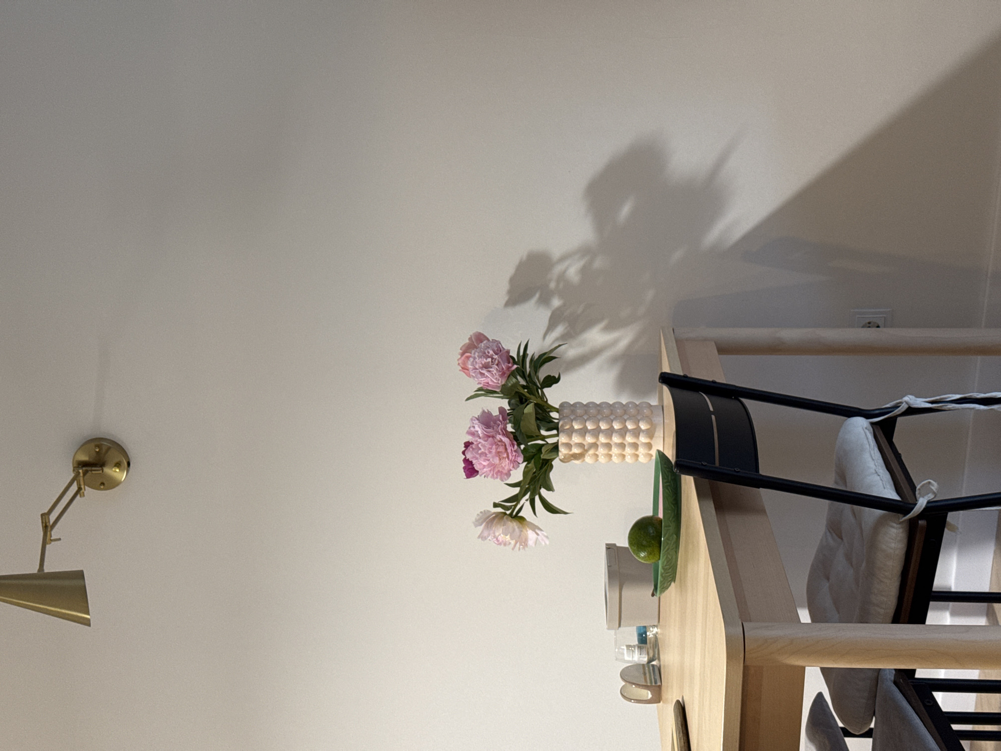 The image shows a simple dining table with a vase of peonies, a green plate with a lime, and other small decorative items. There are chairs around the table, one of which is occupied by a person. The room is illuminated by natural light and a wall-mounted lamp. The scene is set in Izhevsk, Russia.