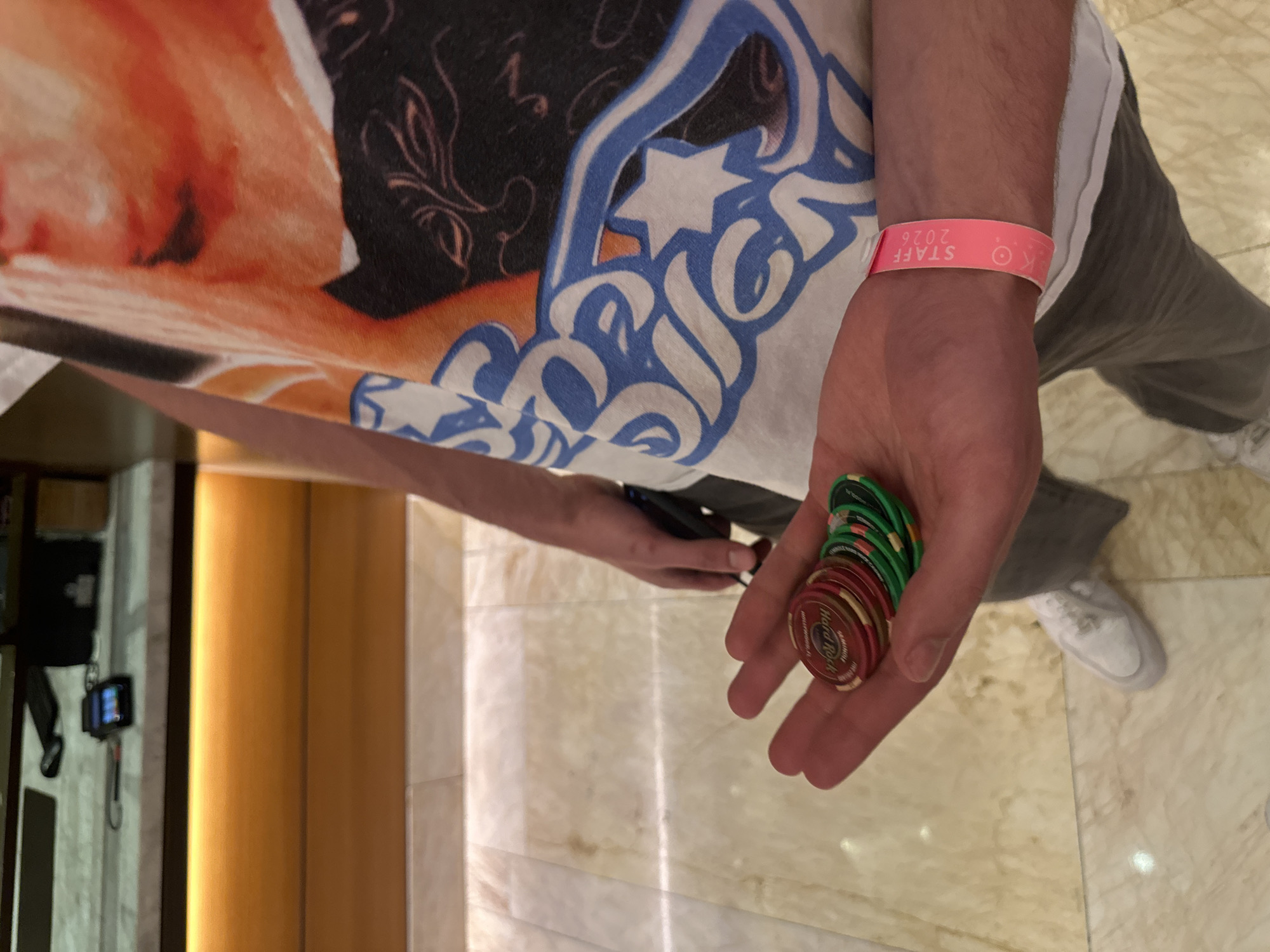 An adult person wearing a t-shirt and holding a stack of Hard Rock casino chips in their left hand and a pink wristband labeled "STAFF 2026". The person is standing on a polished floor, and white tennis shoes are visible near the bottom right of the frame. The overall location is likely the Hard Rock casino in the city of Fort Lauderdale, United States of America.