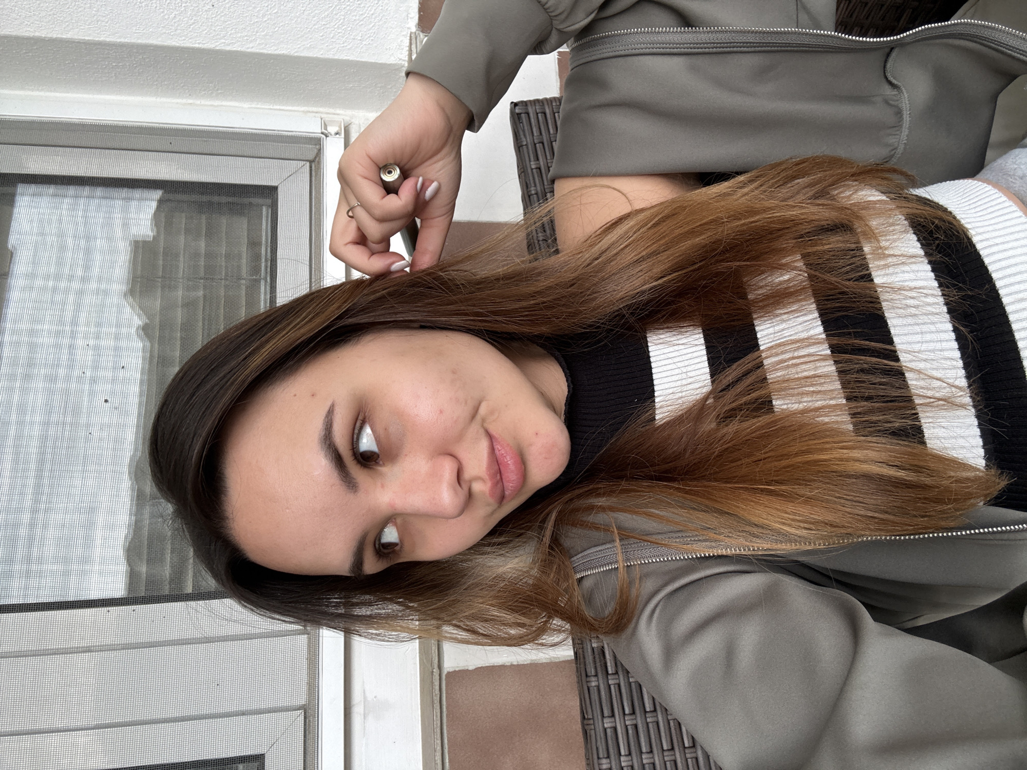 A woman is seated outdoors. She has shoulder-length brown hair with blonde highlights, and is wearing a black and white striped shirt and an olive green jacket. In her left hand, she holds a small metallic object. The background includes a window with a screen, beige tiles, and what appears to be outdoor wicker furniture. Without additional information, it is not possible to confirm the exact location in the city of Sofia, Bulgaria.