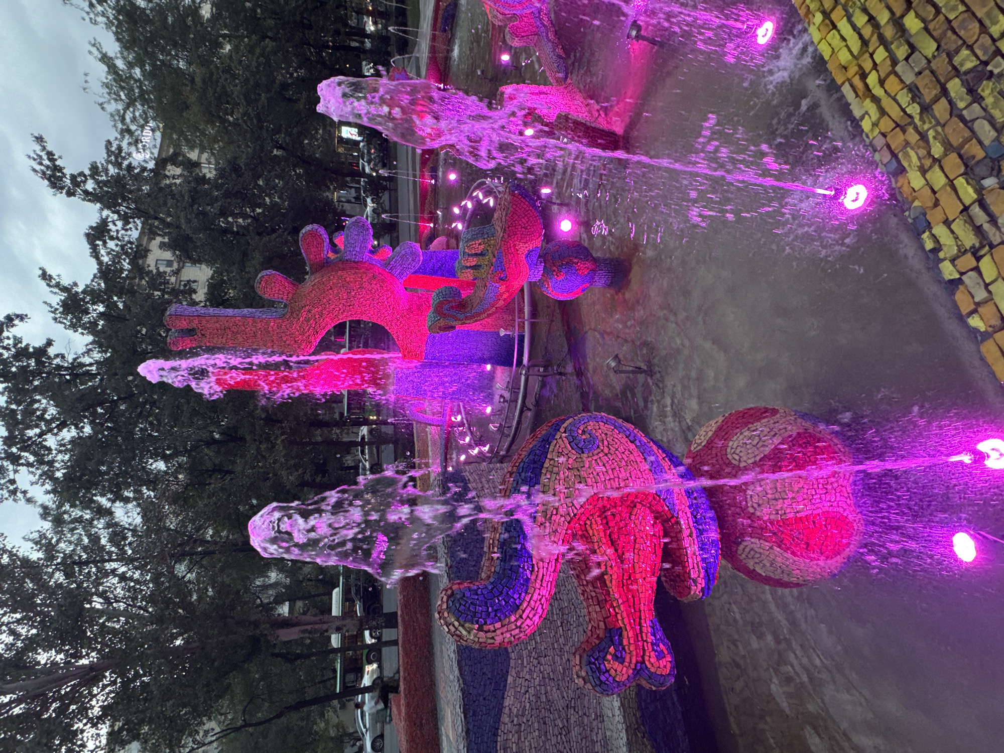 A mosaic fountain is illuminated by pink and purple lights in Bishkek, Kyrgyzstan. The fountain features abstract sculptures covered in colorful mosaic tiles, resembling a stylized fish or dragon-like creature, a hand-like form, and other organic shapes. Water sprays from various points, catching the colored lights and creating a dynamic display. The fountain is situated in a shallow pool, and the surrounding area is paved with mosaic tiles. In the background, lush green trees line a street with cars and a bus, suggesting an urban setting. The sky is overcast, indicating it might be evening or a cloudy day.