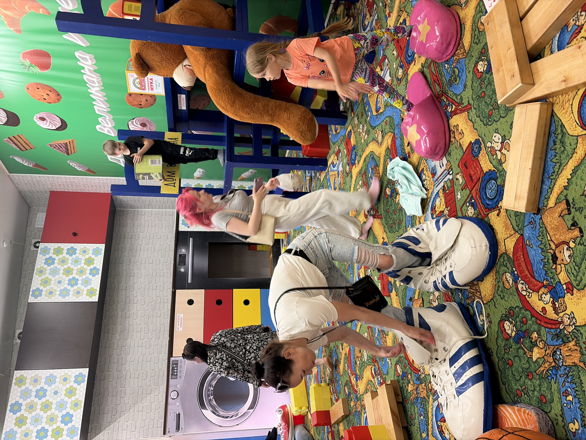 The image depicts an indoor play area, likely a children's entertainment center, with a vibrant and cartoonish carpet featuring farm animals and children. In the foreground, a woman wearing ripped jeans and a white t-shirt is kneeling and tending to two oversized, inflatable Adidas sneakers on the floor. To her right, a young girl with blonde hair, wearing an orange t-shirt and colorful leggings, is standing with her hands out, possibly playing or interacting. Behind the woman, a young boy in a black t-shirt and pants is holding a cardboard box. A large brown teddy bear is visible behind the boy, sitting on blue playground equipment. In the background, a washing machine and other kitchen appliances are depicted on a wall, possibly as part of the play area's theme. The overall scene suggests a playful and engaging environment for children. The specific location in Izhevsk, Russia, is not discernible from the visual information provided in the image.
