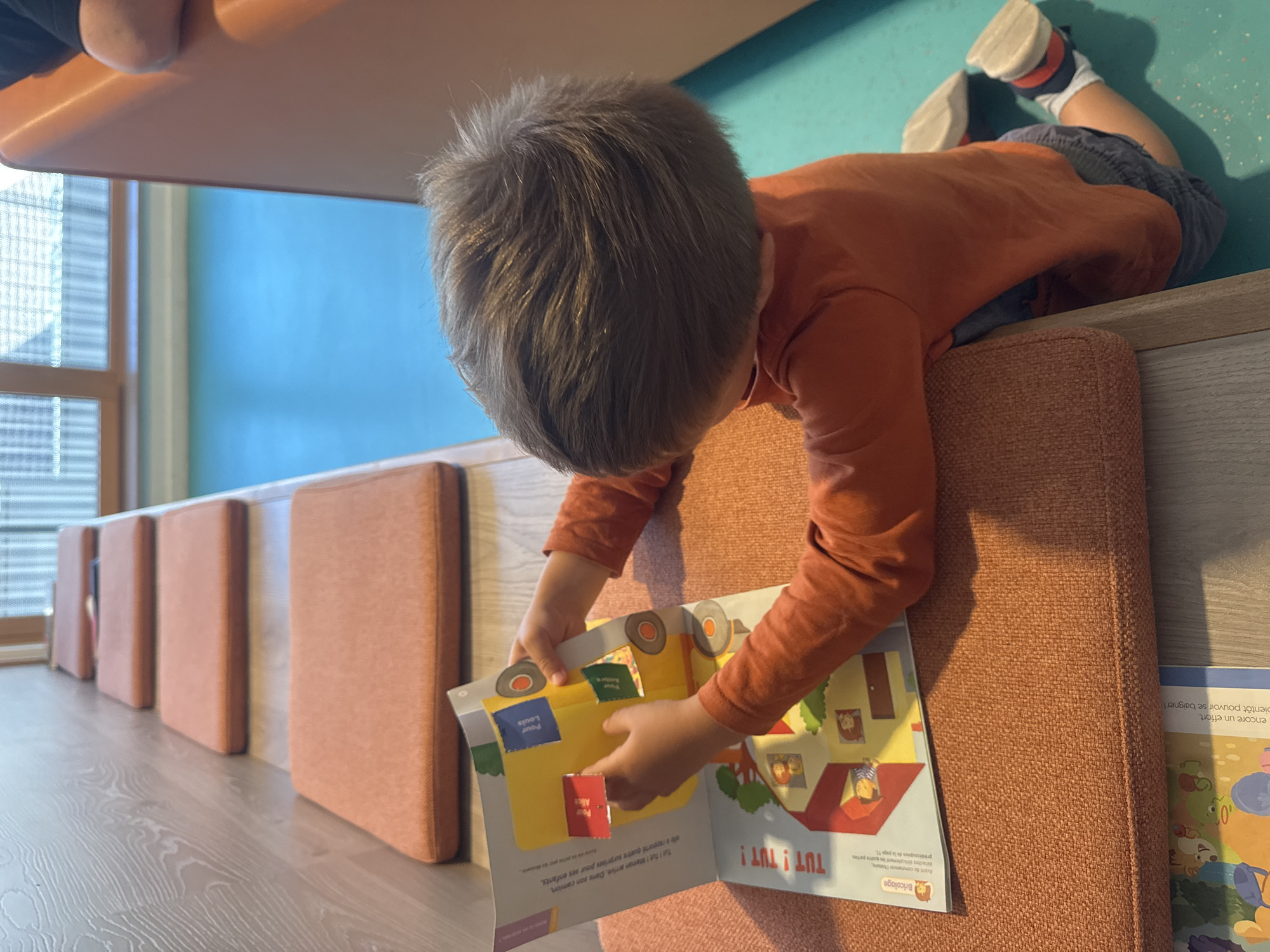 A young child wearing an orange long-sleeved shirt and grey shorts sits on an orange cushioned bench, facing away from the camera. The child is interacting with an open pop-up book that depicts a yellow vehicle and a house with people inside. The book is open on a surface that appears to be another orange cushioned bench. The environment includes wooden paneling on the left and a bright blue floor. The background shows windows with a grid pattern. There is no identifiable information in the image to determine a specific location in the city of Saint-Claude, France.