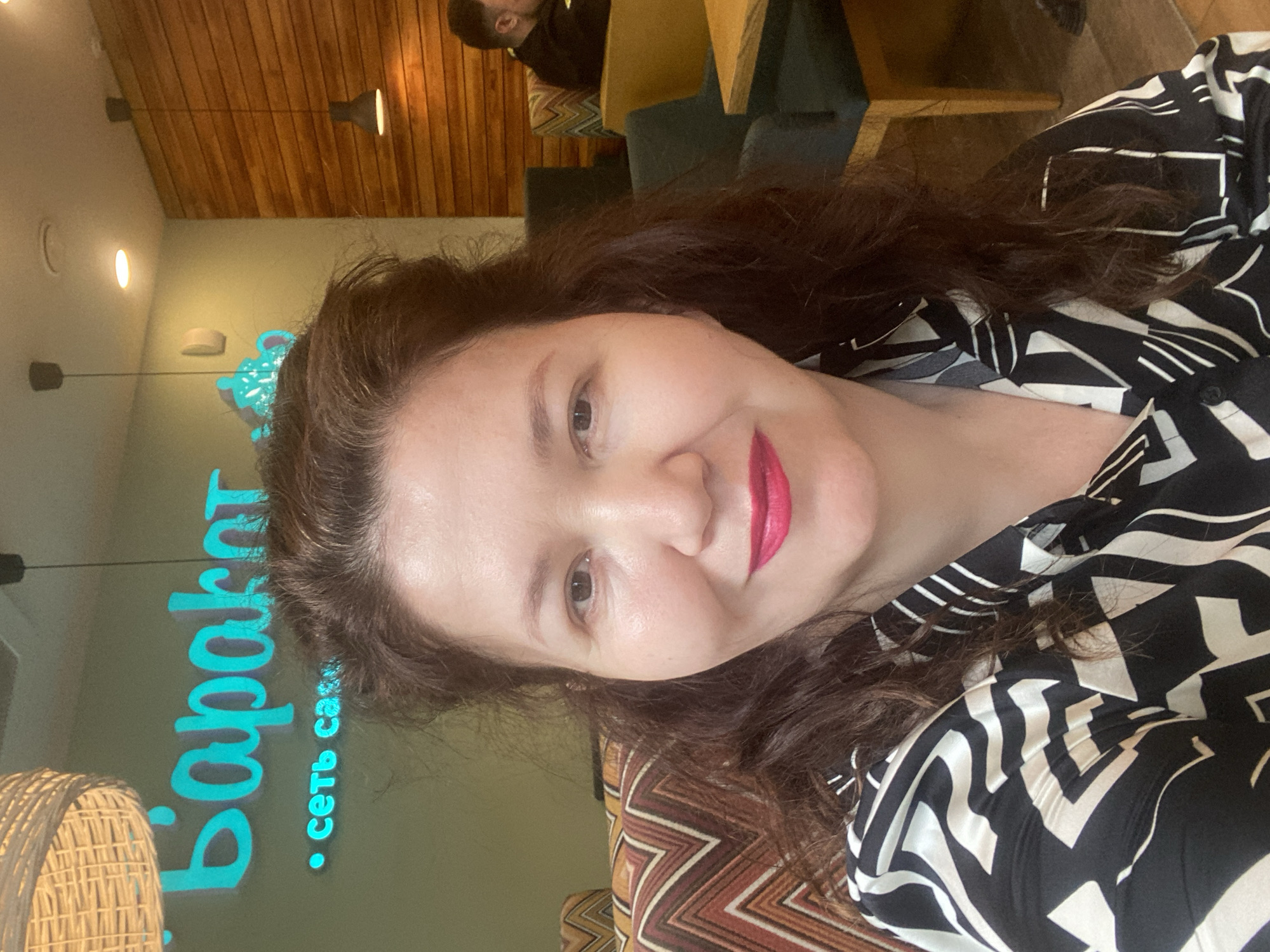 The image is a selfie taken indoors, likely in a cafe or restaurant. A woman with dark, wavy hair and bright pink lipstick is the primary subject. She is wearing a black and white patterned shirt. In the background, a neon sign with Cyrillic text is visible, which reads "Баракет" and "сеть са..." (partially obscured). The wall behind the sign is painted a light green, and further back, a wooden panel wall and hanging light fixtures are visible. In the right side of the background, a man in a dark jacket is seated at a table. The overall lighting suggests an indoor setting with artificial light.
