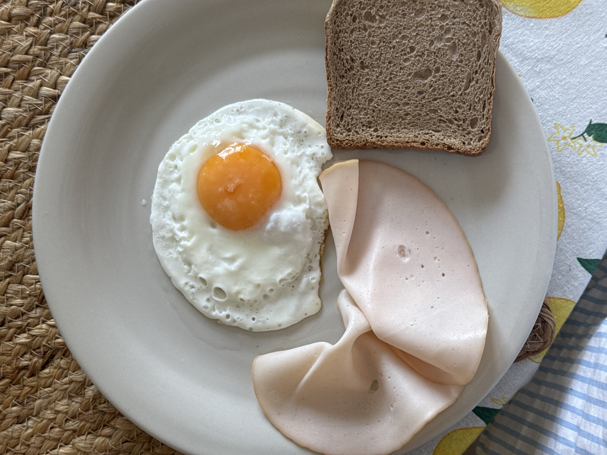A plate of food is presented on a surface. The plate contains a fried egg, a slice of dark bread, and several slices of processed meat. The table is covered with a patterned cloth. A portion of a woven mat is also visible in the frame. The location, Vozdovac Urban Municipality, Serbia, cannot be confirmed from the visual information.
