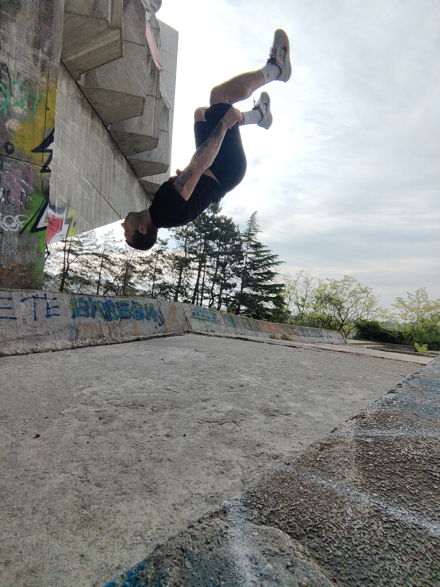 The image captures a man performing a backflip near the Monument of the Bulgarian-Soviet Friendship in Varna, Bulgaria. The man is wearing black shorts, a black t-shirt, white socks, and athletic shoes. He is positioned upside down mid-backflip against a backdrop of a cloudy sky and trees. To the left of the man is a large concrete structure covered with graffiti. Below the man, a concrete platform runs horizontally across the frame. The platform is also covered in graffiti.