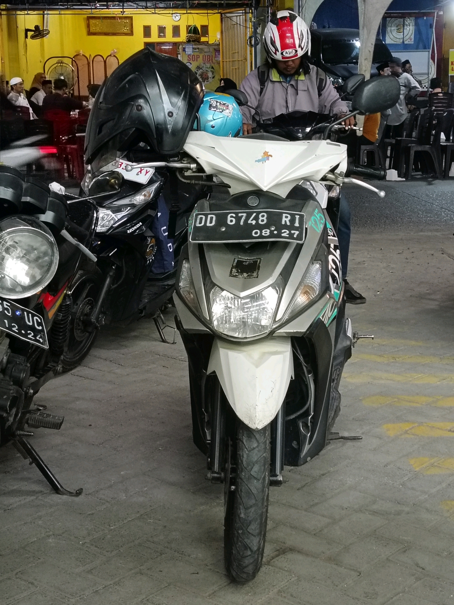 The image captures a street scene in Makassar, Indonesia, with a focus on several parked motorcycles and individuals. In the foreground, a light-colored scooter is prominently displayed, featuring a license plate that reads "DD 6748 RI 08-27". A person wearing a grey jacket and a red and white helmet is seated on this scooter, with their hands resting on the handlebars. To the left of this scooter, another black motorcycle is visible, with a prominent black helmet placed on its front. The background reveals a bustling environment with several people seated at tables, suggesting a food establishment or gathering. The architecture suggests a covered outdoor area, possibly a warung or a small restaurant, with yellow walls and overhead lighting. The ground is paved with grey interlocking bricks. The overall impression is a snapshot of daily life in a commercial or public space in Makassar.