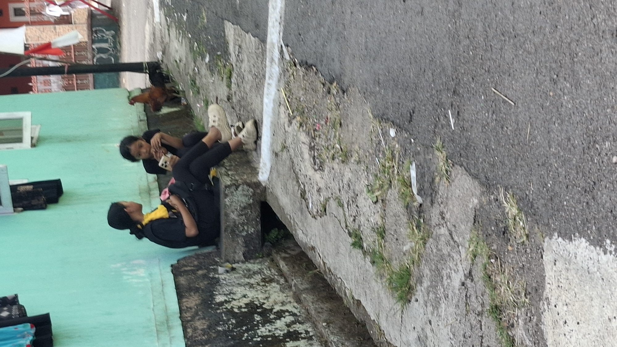 Two young females are seated on a concrete ledge in front of a teal-colored building. The female on the left, wearing a black t-shirt and black leggings, has yellow fabric draped around her neck and is holding a pink item. The female on the right, also in a black t-shirt and black leggings, is looking down at a smartphone in her hands. Both are wearing light-colored sneakers. To the right of the females, a rooster is standing on the ground. The ground in front of them is paved with a cracked concrete surface that transitions into asphalt. Green grass and some debris are scattered on the ground. In the background, a building with red and white Indonesian flags is visible. The scene appears to be an outdoor area in a residential or public space.