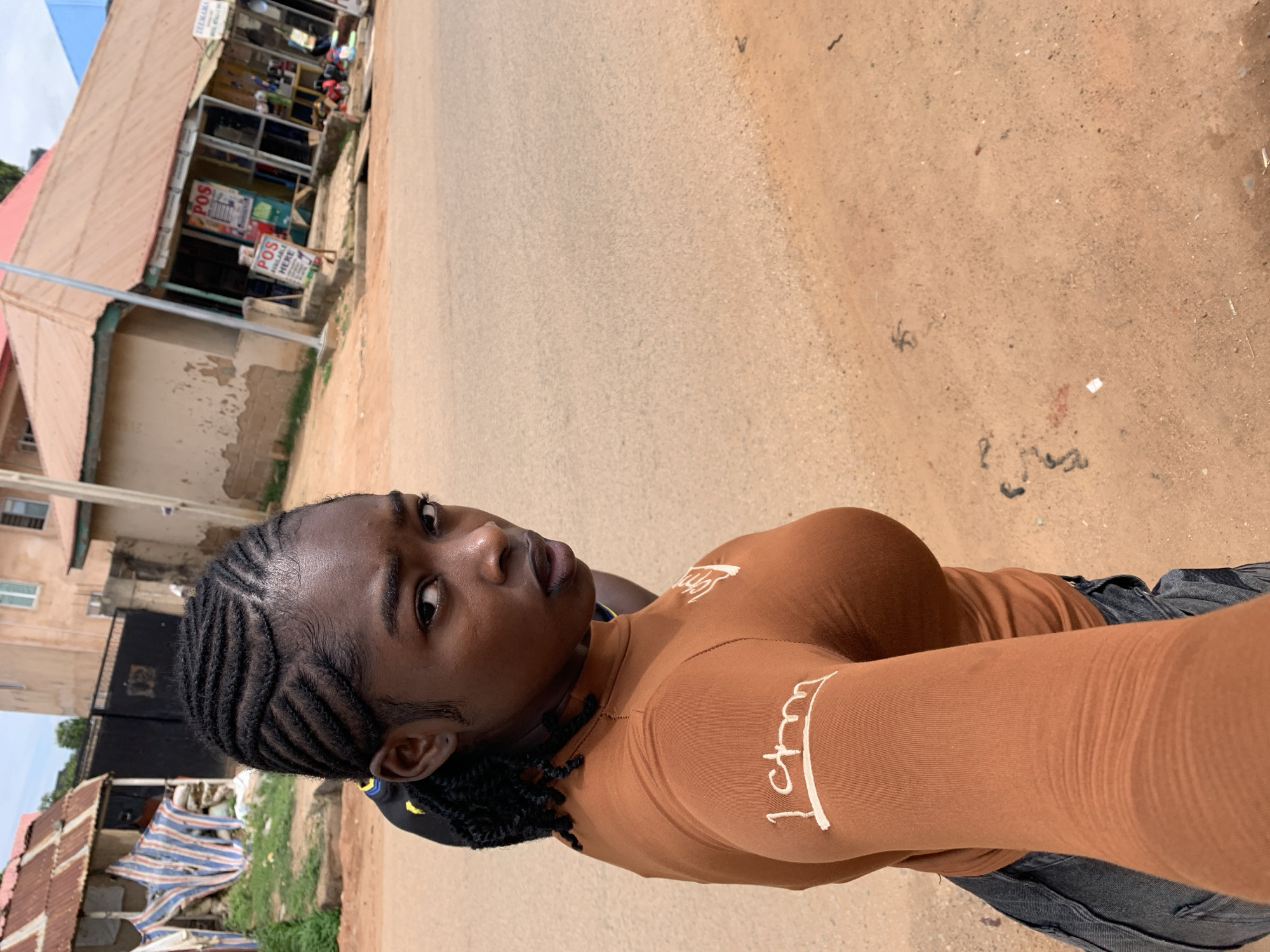 The image depicts a young Black female individual in a street environment in New Stadium, Nigeria. She is positioned in the left foreground, looking directly back towards the camera with a direct gaze and slightly parted lips. Her hair is styled in distinct cornrows, intricately braided across her scalp. She wears a form-fitting, long-sleeved, rust-brown top with white stylized lettering, resembling "1ctm," visible on her left sleeve. Dark-colored pants or jeans are partially visible below her waist. A segment of a yellow detail on a dark object is discernible behind her left shoulder, possibly indicating another person or item carried.
The immediate ground around her consists of reddish-brown dirt, with scattered small debris. To her left background, a dilapidated structure with a rusty corrugated iron roof and exposed, weathered walls stands alongside a tattered, striped fabric shelter. Further back and to the right of this structure, a black metal gate is visible.
The central and right portions of the image feature a light grey asphalt road, transitioning to a reddish-brown dirt shoulder on the far right. Along the right side of the road, a commercial strip is visible, comprising several single-story buildings with corrugated iron roofs. One prominent building has a reddish-brown roof and cream-colored walls. In front of this building, a red sign with "POS AVAILABLE HERE" in white lettering is clearly displayed, alongside other less legible signs. A tall, slender utility pole stands on the right side of the road, adjacent to the commercial structures. The sky above is light and hazy.