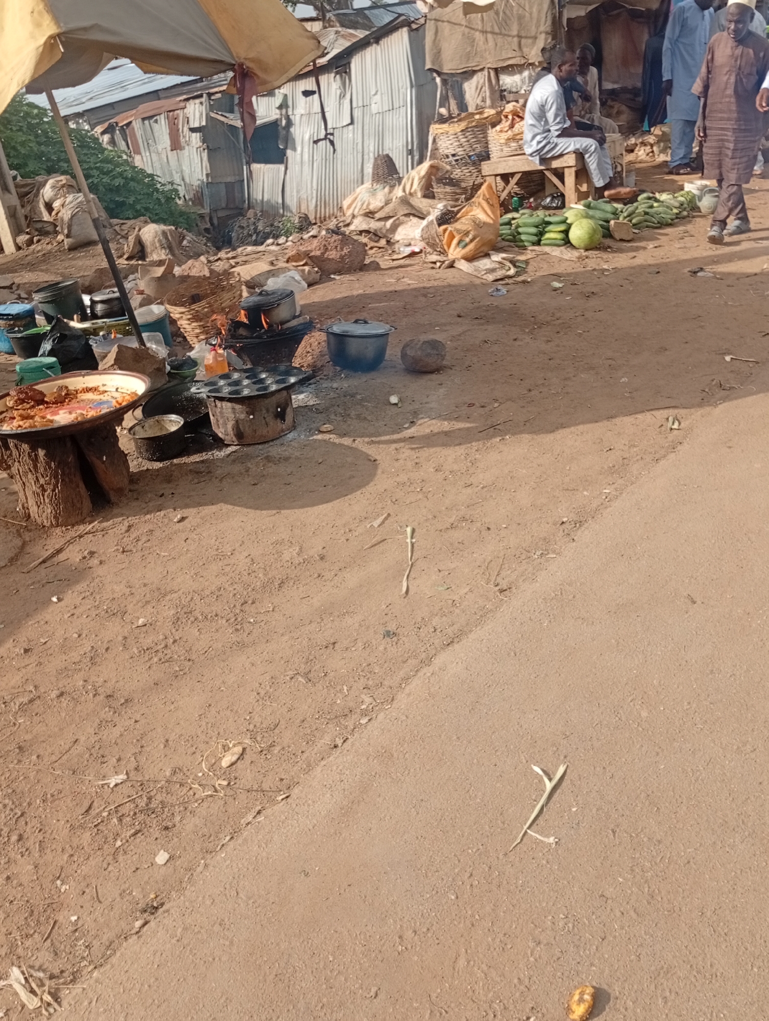 The image depicts an outdoor market scene in Rafin Par, Nigeria. In the foreground, the ground is sandy and dusty, with scattered debris including dry leaves and small stones. A paved road or path runs diagonally across the bottom right of the frame. In the midground and background, there is a collection of market stalls and vendors. Several individuals are present, including a man in a brown traditional outfit walking towards the viewer from the right. To the left of the frame, a makeshift stall is set up under a large, weathered yellow umbrella. Various cooking pots and utensils are arranged on the ground and on wooden stands, some with food being prepared. There is a prominent round, dark object on the ground to the left of the cooking area, possibly a large stone or coconut. Behind the stalls, a corrugated metal structure forms the backdrop, appearing to be part of makeshift shelters or buildings. A pile of green produce, including cucumbers and a large melon, is displayed for sale on the ground to the right of center. A man in a light-colored garment is seated on a wooden bench amongst the produce. The overall atmosphere suggests a busy, informal marketplace.