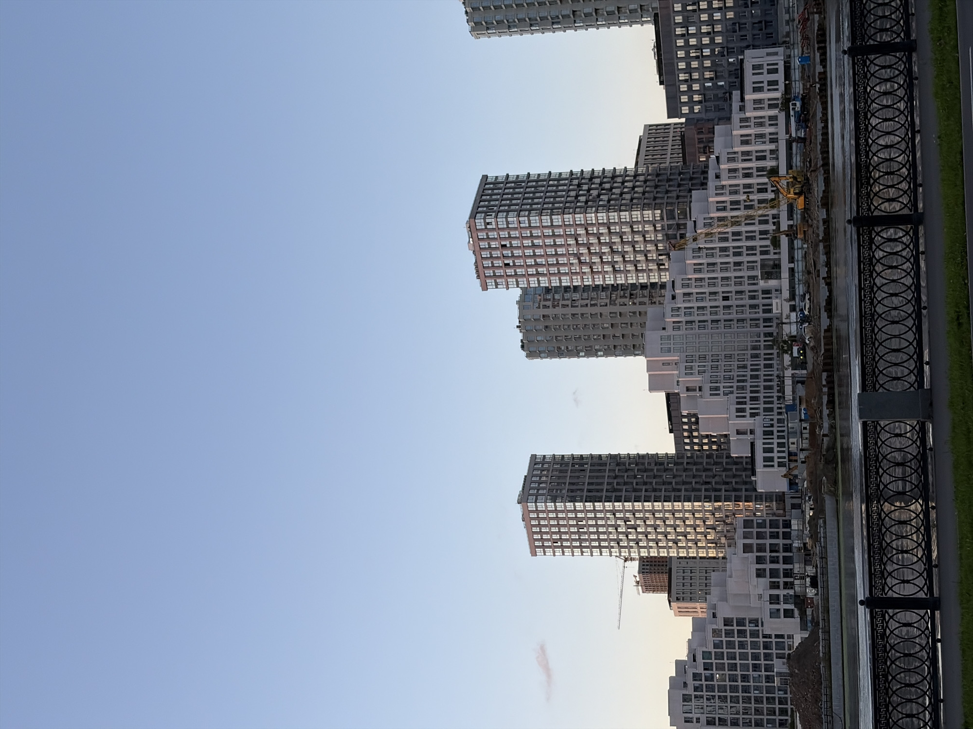 The image depicts a modern cityscape with a clear blue sky. The foreground features a black metal fence with an intricate oval pattern, bordering a grassy area. Beyond the fence, a calm body of water reflects the sky and the surrounding buildings.
The middle ground is dominated by a complex of contemporary residential buildings. These buildings vary in height and architectural style, with many featuring large glass windows that reflect the light. Some of the buildings are tall, skyscraper-like structures, while others are lower-rise complexes. A construction crane is visible among the buildings, suggesting ongoing development in the area. Various construction vehicles, including an excavator and trucks, are present on the construction site.
In the background, more tall buildings and structures are visible, contributing to the dense urban environment. The overall scene suggests a developing waterfront district in a city, with a mix of completed and under-construction residential and commercial properties. Given the modern architecture and the presence of a crane, this scene is likely located in a rapidly developing area of Moscow, Russia, possibly a new residential complex being built along a river or canal.