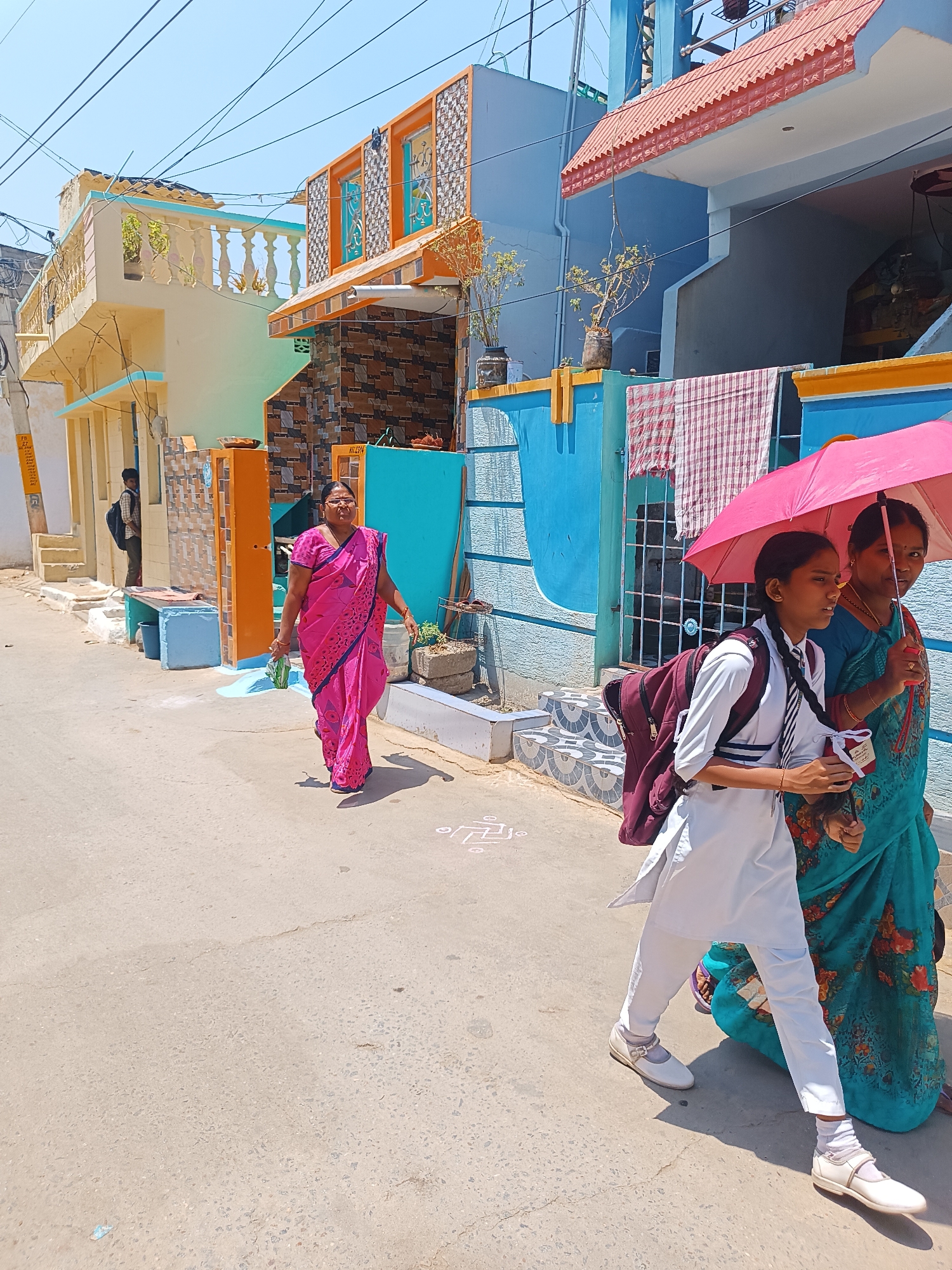 A daytime street scene in Pedda Kammavaripalli, India, features pedestrians on a paved thoroughfare bordered by residential buildings. In the foreground-right, a young female, identified as a student by her white uniform (white shirt, pants, black-and-white tie), white shoes, and maroon backpack, walks alongside an adult female in a teal floral saree. The adult holds a bright pink umbrella aloft, shading both individuals. The student holds a small object in her right hand; her hair is styled in two braids. Towards the center-left, an adult female in a bright pink floral saree, wearing glasses and carrying a small green bag, walks in the opposite direction. In the distant left background, a male figure with a dark backpack is partially visible. The surrounding environment consists of multi-storied houses painted in distinct colors such as light blue, yellow, and orange, featuring balconies with balusters, patterned wall tiles, and exposed overhead utility lines against a clear sky. Strong shadows indicate direct sunlight. A white traditional "kolam" pattern is visible on the pavement surface on the right.