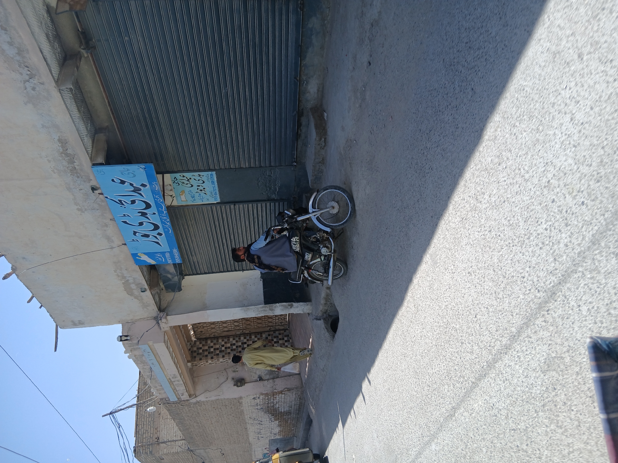 This image depicts a sunlit street scene in Quetta City Tehsil, Pakistan, captured from a slightly tilted, elevated perspective. The setting appears to be an urban or semi-urban environment with buildings lining one side of the road.
In the mid-ground, a person wearing a dark shirt and an orange cap is seated on a black motorcycle, which is parked parallel to a building. The building features a large, grey metal rolling shutter, indicating a closed shop. To the left of the shutter, a vibrant blue rectangular sign is affixed to the building's facade. Further down the street, another individual, dressed in a light-colored traditional outfit (likely shalwar kameez), is visible either walking or standing near an entrance. The street surface is paved, possibly concrete or asphalt, and a distinct diagonal shadow from an unseen structure falls across it, signifying bright daylight conditions and a clear sky. A small, dark drain or hole is present in the road near the motorcycle.
The prominent blue sign displays text in Urdu/Persian script. The visible text reads "حاجی عبادی بادی و جواد" (Haji Abadi Badi o Jawad), which is likely the name of the business or its proprietors. Below this, an address is provided: "پتہ - آخری سناپ نزد گول مسیر کوئٹہ" (Address - Akhri Snap near Gol Maseer Quetta). Two phone numbers are also discernible: "0513-8671775" and "050-771106". Additional text, "جارتی خبر استار ارمیان منی," is also present, possibly indicating a service or slogan. The overall scene suggests a typical street with local businesses in a South Asian city.