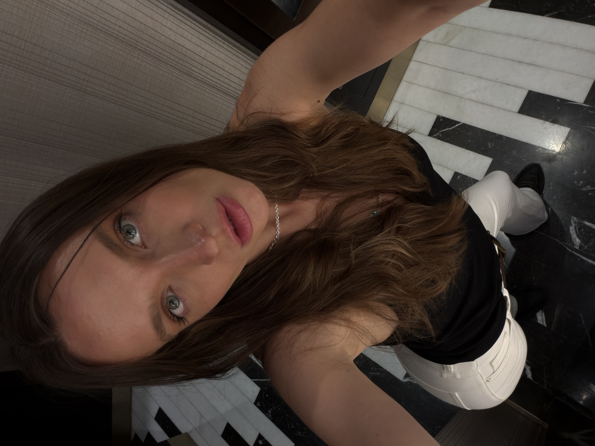 A woman takes a selfie in an indoor setting, likely the Avala Hotel in Budva, Montenegro, based on the distinctive black and white striped floor. The woman has long brown hair, fair skin, and is wearing a black top and white pants. She is also wearing a delicate silver necklace. The background consists of a black and white geometric floor pattern and a glimpse of a dark-colored wall.