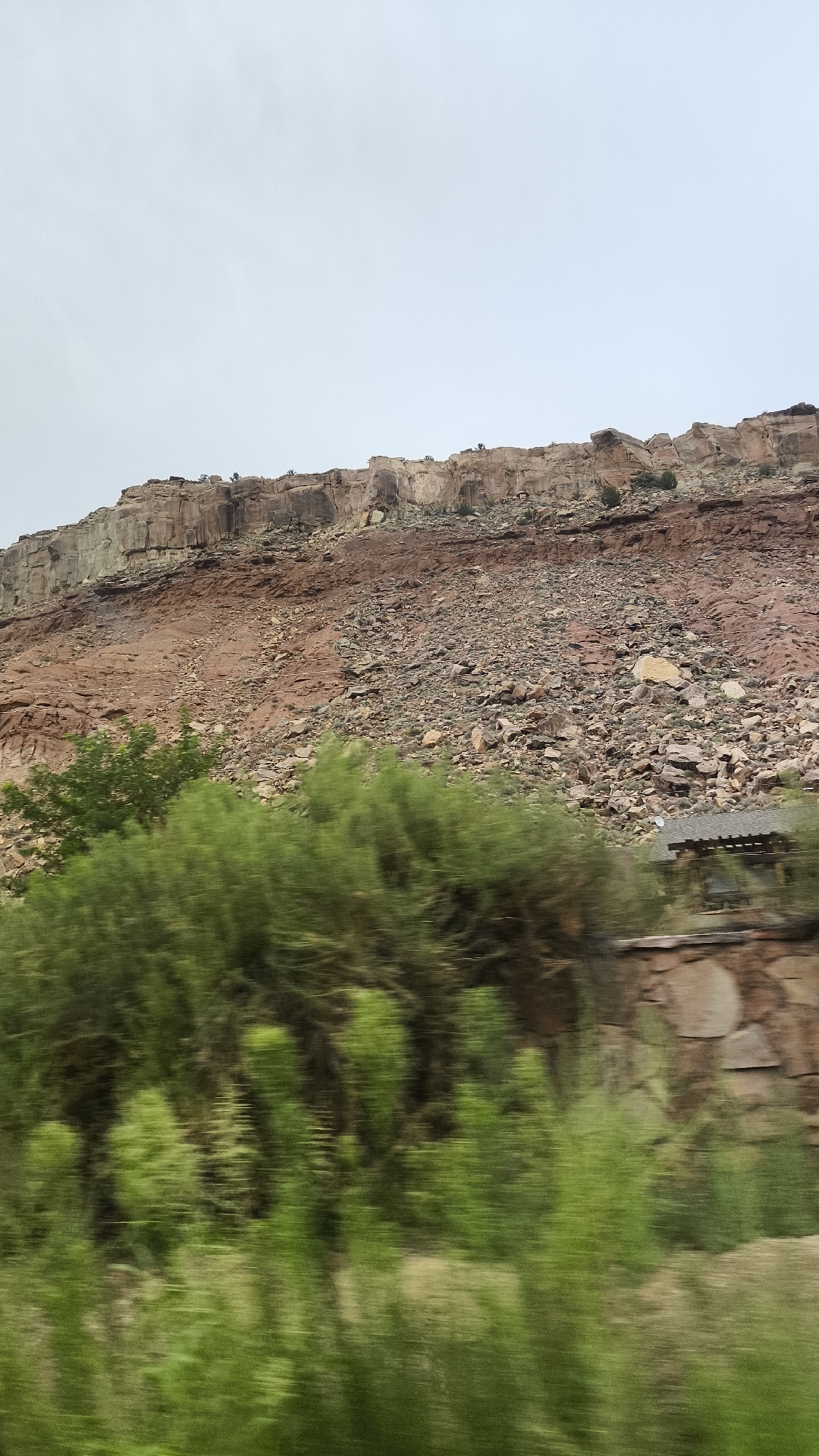 The image shows a blurred landscape of a rocky cliff face under a pale sky. The cliff has distinct layers of red and tan rock, with a scree slope of smaller rocks and debris at its base. In the foreground, out-of-focus green foliage, likely trees and bushes, obscures the lower part of the scene. Partially visible behind the foliage on the right side of the image is a stone structure, possibly a building or wall, with a dark roof. The motion blur suggests the image was taken from a moving vehicle. The scene is situated in Rockville, United States of America.