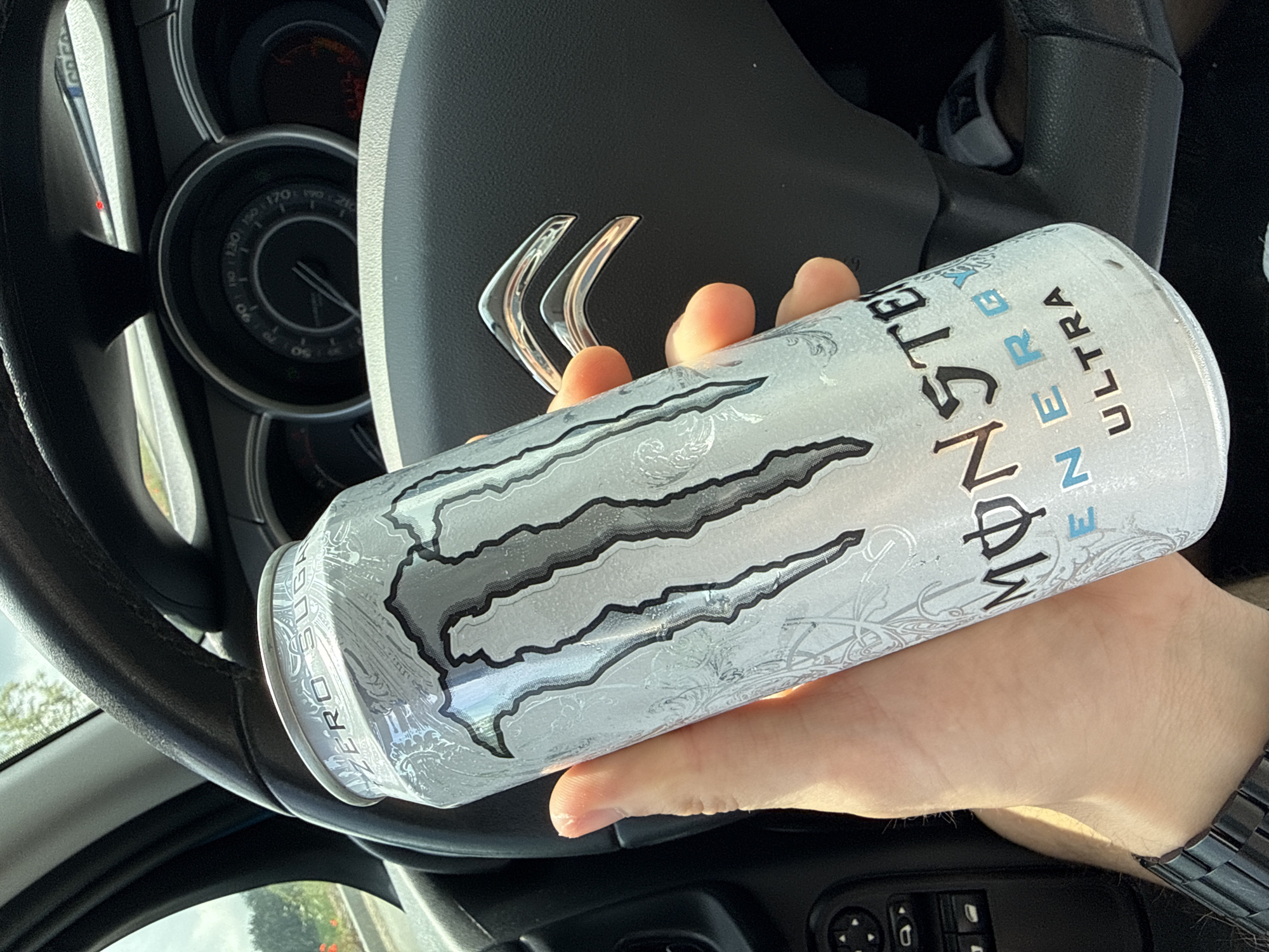 The image captures a close-up view of a person's hand holding a silver Monster Energy Ultra energy drink can within a car interior. The can prominently displays the black and white Monster M-claw logo, along with the text "MONSTER ULTRA", "ENERGY", and "ZERO SUGAR".
The setting is clearly inside a vehicle, likely from the driver's perspective. A black, leather-wrapped steering wheel is visible, featuring a chrome emblem composed of two stylized, layered chevron shapes, which is characteristic of a Citroën car. Behind the steering wheel, a portion of the dashboard's instrument cluster can be seen, including an analog dial with numerical markings indicating speeds up to at least 210 km/h. Below the person's hand, part of a car door panel with control buttons for windows and possibly mirrors is visible.
The scene is well-lit by bright daylight, suggesting the photograph was taken during the day. The hand appears to be an adult's, and a metallic watch is worn on the wrist. The activity depicts a casual moment, perhaps while driving or during a stop, in Ruen, Bulgaria.