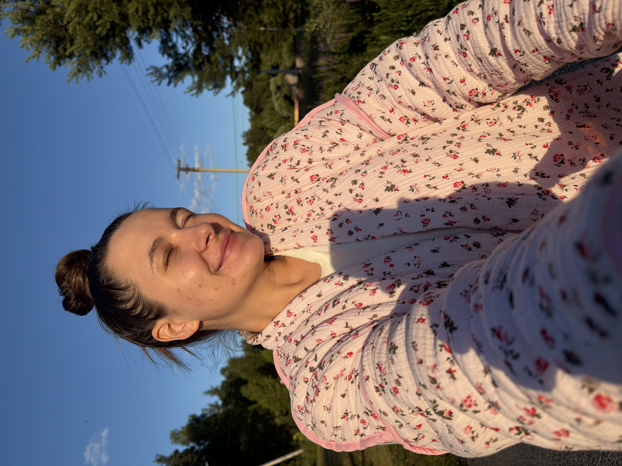 The image shows a young woman outdoors, leaning back and closing her eyes as if enjoying the sun. She is wearing a light pink, floral-patterned robe. Her hair is pulled back into a bun. The background consists of a clear blue sky, green trees, and a utility pole with wires. The lighting suggests it is either early morning or late afternoon, with the sun casting a warm glow on her face and robe. The scene appears peaceful and relaxed. No text is visible in the image.