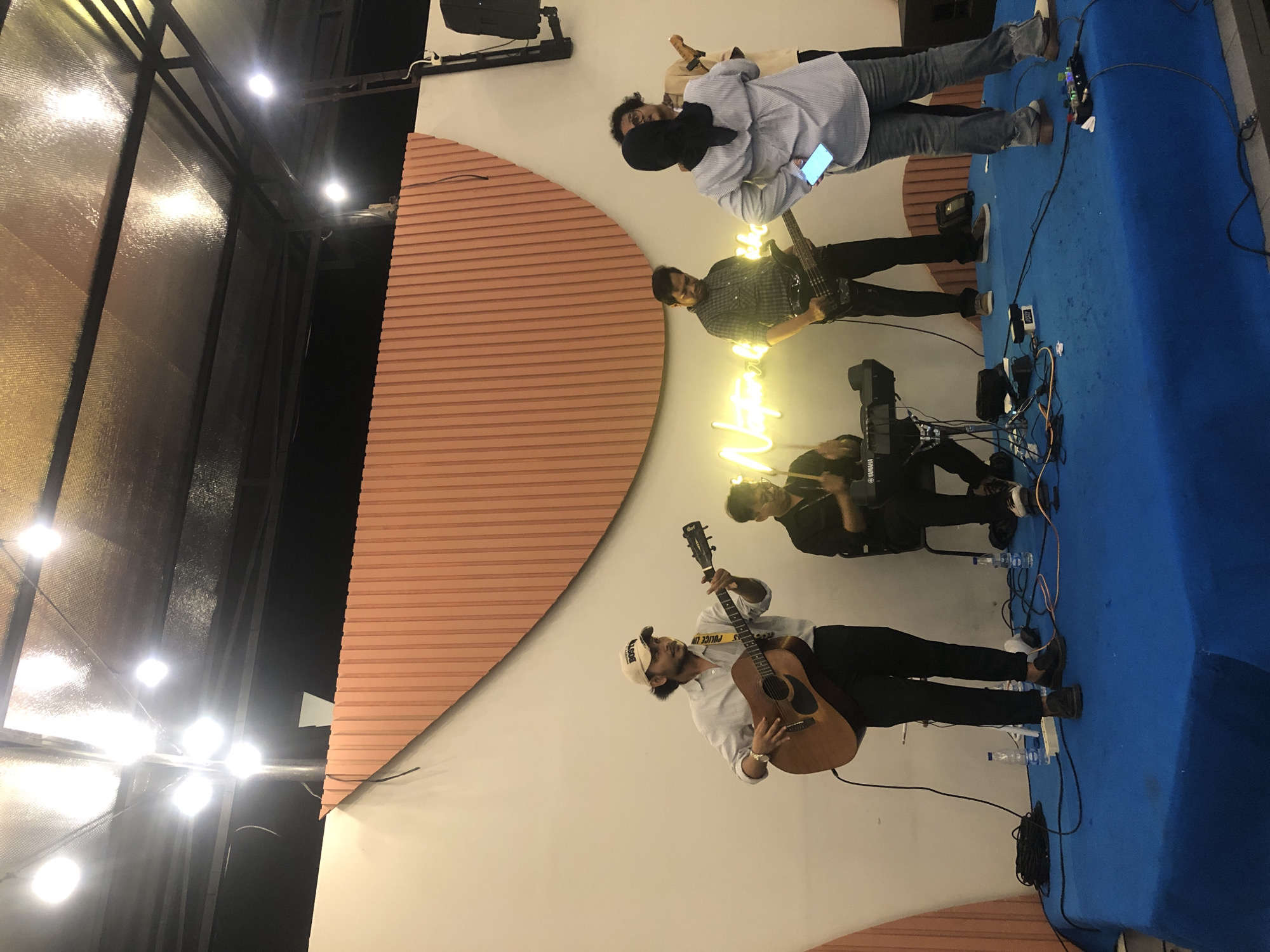 The image shows a band of five musicians performing on a blue carpeted stage in Lhokseumawe, Indonesia. The stage is against a white wall decorated with a large semicircle of horizontal orange lines. From left to right, the musicians are: a woman in a light blue shirt and hijab looking at her phone; a man in a black and white plaid shirt playing bass guitar; a seated man playing a keyboard; another seated man playing drums; and a man in a white shirt and backwards baseball cap playing an acoustic guitar. The stage has power cables, equipment, and water bottles scattered around the musicians' feet. The room is lit by several bright lights.