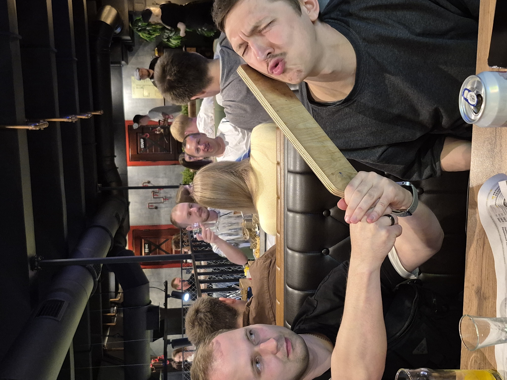 This media file, captured in Izhevsk, Russia, depicts an indoor gathering, likely a restaurant or bar. The scene is filled with people, with several individuals prominently featured. In the foreground, a man with a mustache and closed eyes is leaning against a wooden board, his mouth pursed as if kissing or making a funny face. His expression suggests a playful or lighthearted mood. To his left, another man is visible, his face partially obscured. Further back, a group of people are seated at tables, some holding drinks and interacting.
The setting appears to be dimly lit, creating a cozy atmosphere. Exposed pipes and industrial-style decor are visible, hinting at a modern or trendy establishment. A can of beer is placed on a wooden surface near the foreground subject. The people are dressed casually, suggesting a social outing. The overall impression is one of friends enjoying each other's company in a relaxed environment. The time of day is indeterminate due to the indoor lighting, but the social gathering implies it's likely evening or nighttime. No specific weather conditions can be inferred. There is no visible text in the frame.