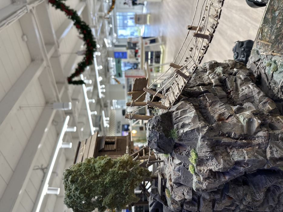 This media file captures a detailed miniature model or diorama, presented from a low-angle perspective. The foreground features a rugged, mountainous landscape composed of dark grey and brown rocky formations with visible stratification and sparse green moss. Spanning across a gap in the rocks is a miniature, rickety suspension bridge made of wooden planks and ropes, suggesting age and wear. To the left, a small, rustic wooden building or shack is nestled amidst miniature green trees or shrubs, appearing to be integrated into the terrain. On the far right, near the top of a rock formation, a small, dark object resembling a miniature camera or part of the model is visible.
The background is heavily blurred but indicates a bright, spacious indoor commercial or retail environment. It shows a white ceiling with exposed beams and numerous rectangular light fixtures. A festive red and green garland is draped across the ceiling, suggesting holiday decorations. The partial text "SONOS" is faintly visible on a blurred sign or display in the background. The scene is static, showcasing the intricate details of the miniature landscape within this indoor setting. The image is devoid of human subjects, and no specific time of day or weather is discernible from the indoor context. This detailed display is located in Metamorfosi, Greece.