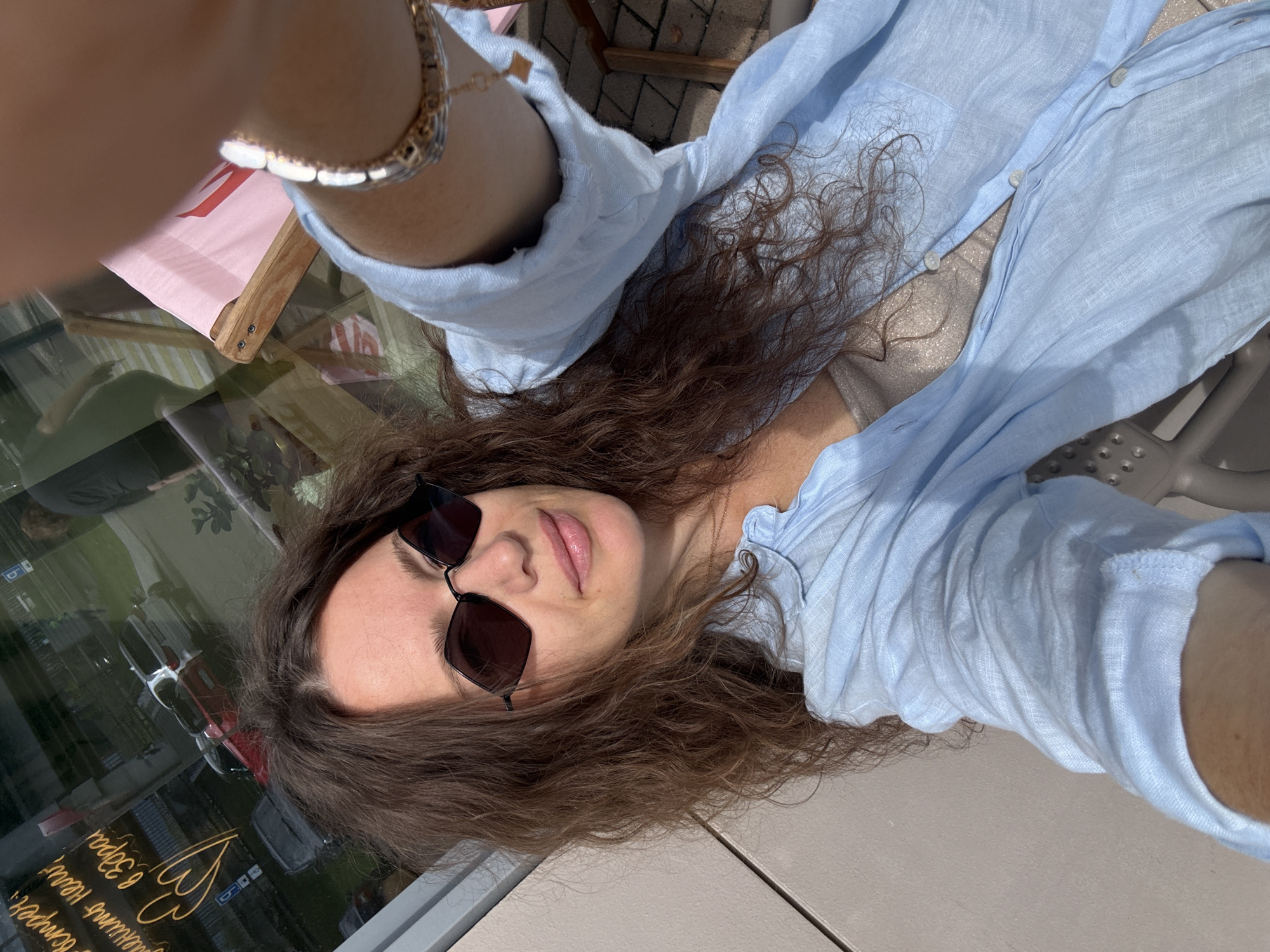 A selfie taken from a low angle shows a woman with long, wavy brown hair and dark-rimmed sunglasses. She is wearing a light blue linen shirt, partially unbuttoned, revealing a shimmery beige tank top underneath. A delicate gold necklace is visible. On her left wrist, she wears a silver bracelet. The background suggests an outdoor setting, possibly a cafe or restaurant patio in Moscow, Russia. Reflected in the glass window behind her are cars, buildings, and a pink deck chair. The overall impression is a casual, sunny day.