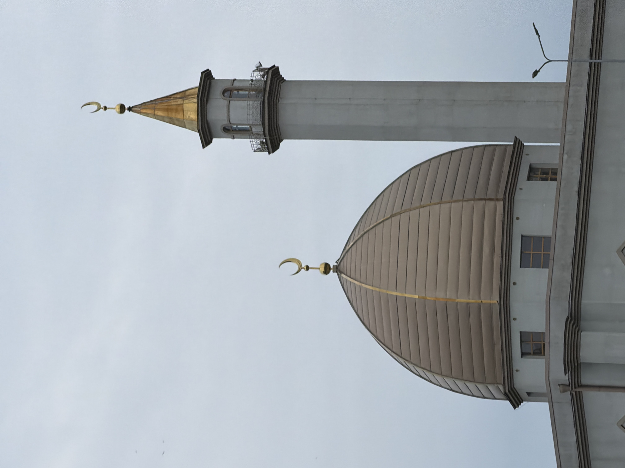 The image shows a low-angle view of a mosque, likely photographed in Saint Petersburg, Russia, given the architectural style prevalent in that region. The mosque features a prominent dome with a crescent moon finial on top. A minaret, also topped with a crescent moon, rises behind the dome. The mosque has a light gray or white facade, and the dome is covered with brown panels. The sky is overcast and gray.