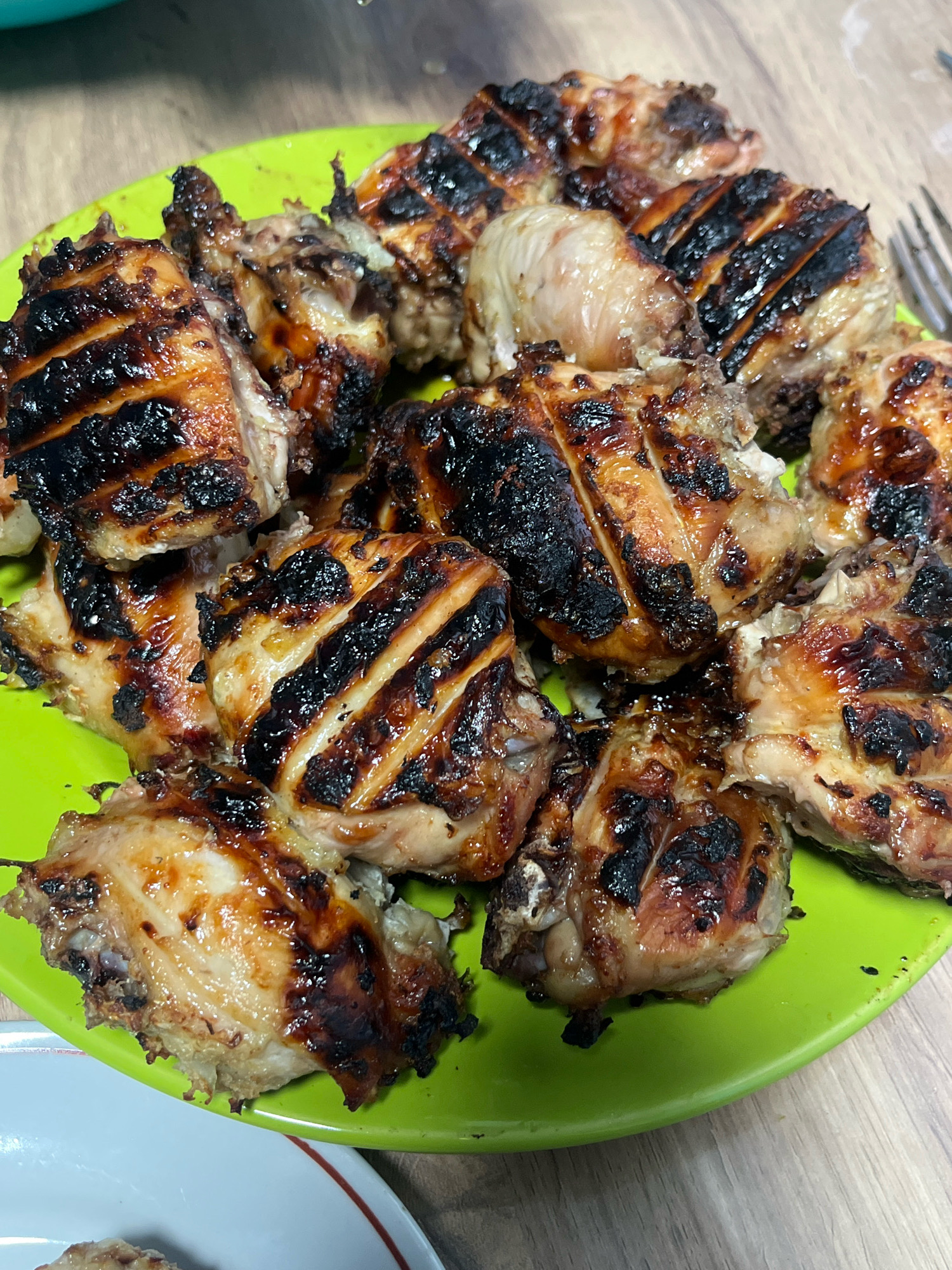 A close-up, high-angle, eye-level shot shows a bright green plate filled with grilled chicken pieces. The chicken is heavily charred in some areas, with visible grill marks across the surface of many pieces. The meat appears to be a mix of light and dark meat, with some pieces showing skin. The plate rests on a wooden surface, possibly a table, and a fork is partially visible on the right side of the frame. The overall lighting suggests an indoor setting. There are no discernible features indicating the specific location within the city of Nizhny Tagil, Russia.