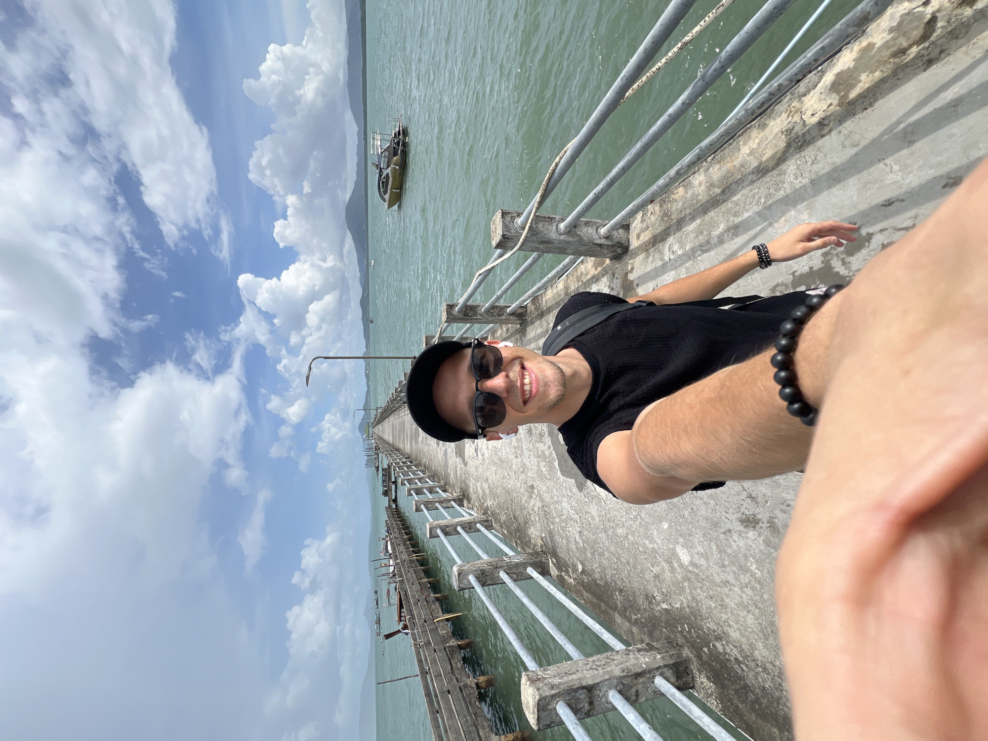 A man, wearing a black t-shirt, black baseball cap, sunglasses, and white earbuds, takes a selfie while standing on a concrete pier. The pier extends into a body of water under a cloudy sky with blue patches. To the left of the pier, a wooden walkway with multiple boats tied to it is visible. In the distance, mountains are seen across the water, and a modern-looking boat is anchored. The man has a black beaded bracelet on his left wrist. The scene appears to be located in Pa Khlok, Thailand.