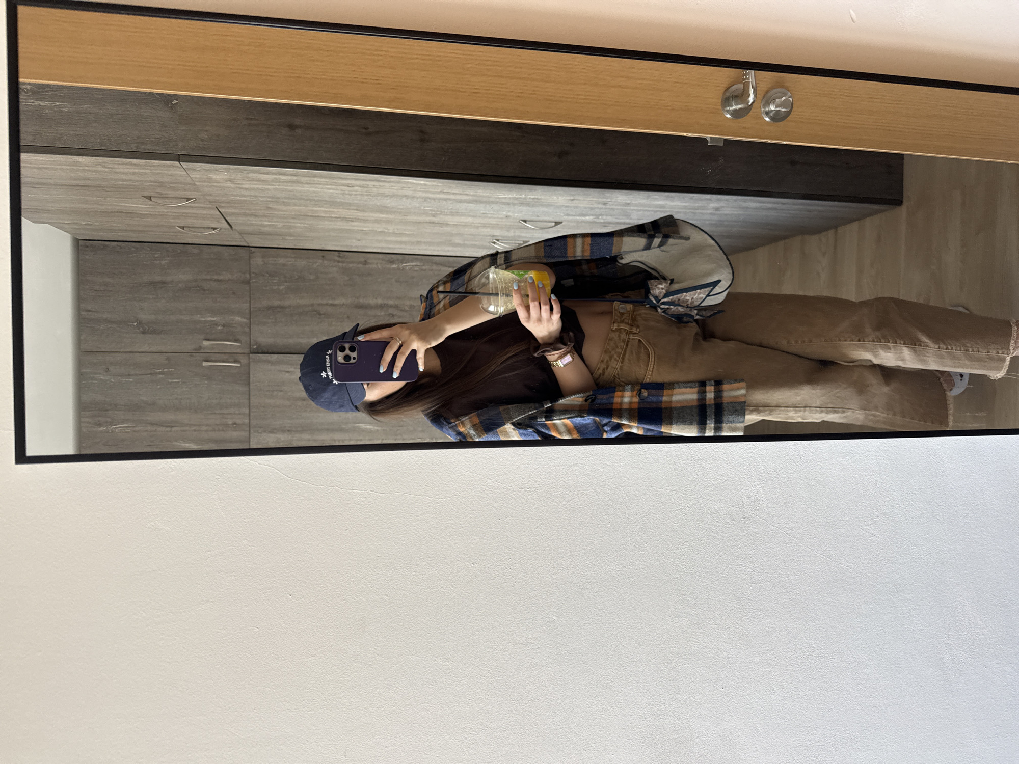 An individual is depicted in a full-length mirror selfie taken indoors. The subject, whose face is obscured by a purple smartphone held in the right hand, is wearing a dark blue baseball cap with white embroidery, a dark brown cropped short-sleeved top revealing the midriff, and light brown wide-leg pants with frayed hems. A blue, orange, and brown plaid button-up shirt is draped open over the shoulders. The left hand holds a clear plastic cup with a lid, straw, and a yellow-orange liquid containing fruit pieces. Accessories include a silver ring, a scrunchie, and a watch on the left wrist, and a small white purse with a patterned scarf hanging from the left shoulder. The reflection reveals a room with a large, dark gray wood-grain wardrobe with silver handles occupying the background. To the far right of the mirror, a light brown wooden door with a silver doorknob and lock mechanism is visible. The floor appears to be a light wood tone. The wall adjacent to the mirror on the left is plain white, and a light pinkish wall is partially visible on the far right, outside the mirror's frame. The scene is an indoor setting, likely a private residence, within the city of Plovdiv, Bulgaria; no specific identifying external features are visible to pinpoint a precise location within the city.