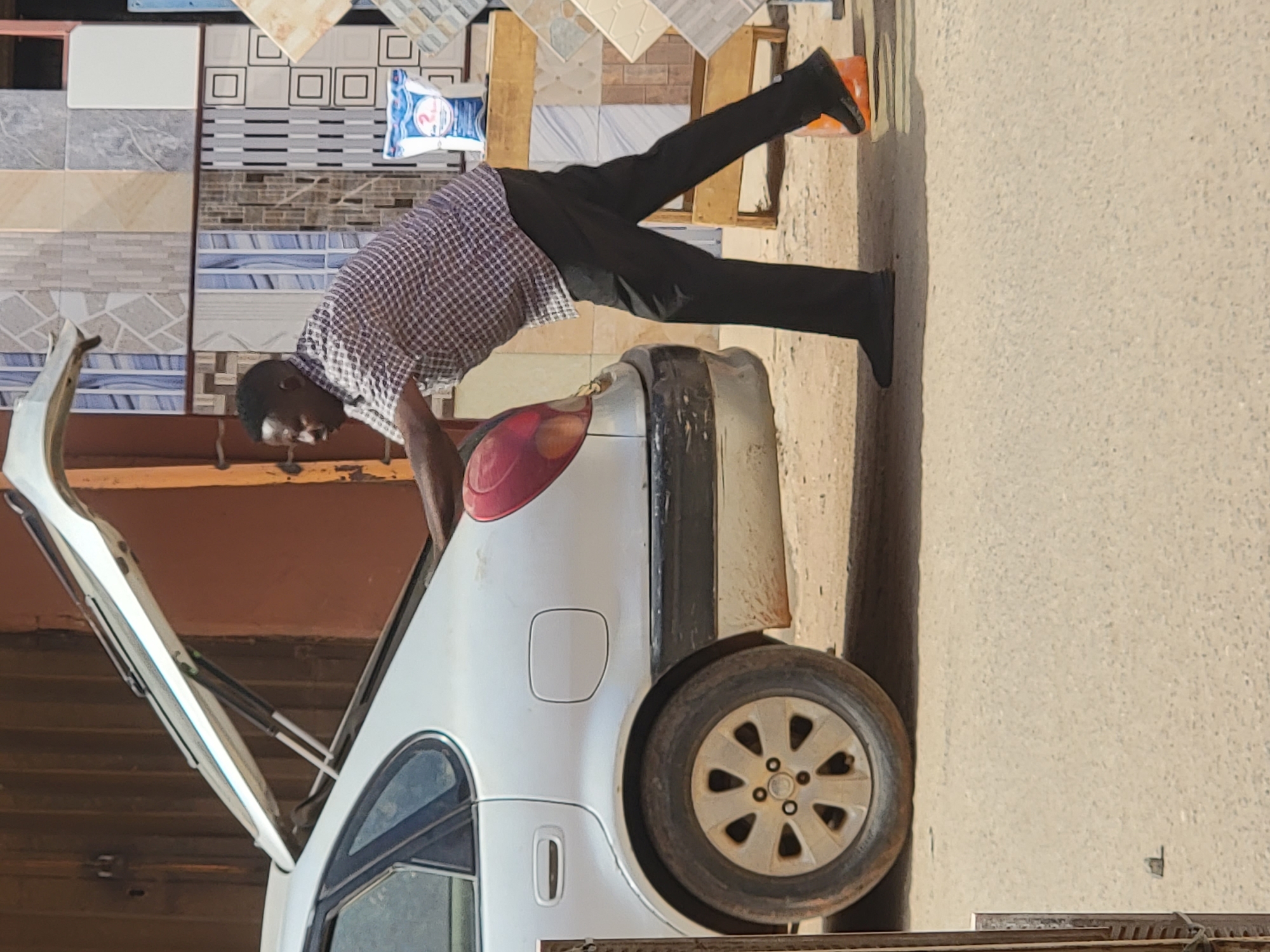 A daytime scene in what appears to be an outdoor commercial area or market in Maiburiji, Nigeria, features a man interacting with a white car. The man, of African descent, wears a checkered shirt, dark trousers, and black shoes. He is captured in the act of leaning into the open trunk of the white car, suggesting he is either loading or unloading items from the vehicle. His posture indicates focus on the task.
The car is a light-colored sedan, visible from its rear and side. Its trunk lid is fully open, revealing the car's interior. Notable details of the car include a red taillight, a fuel cap on the rear fender, and a multi-spoke wheel that appears dusty.
The background behind the man and car is dominated by a display of various floor or wall tiles. These tiles are stacked in different patterns and colors, including white, beige, grey, and blue, with designs ranging from plain to patterned and marbled. They are arranged on what look like wooden pallets or display stands, typical of a tile shop or a roadside vendor. A blue and white bag, possibly containing cement or a similar building material, with some unreadable text, is also visible among the tiles. The ground beneath is a light-colored, dusty, and somewhat uneven surface.
The bright lighting suggests it is daytime, under clear weather conditions. The setting conveys a practical, everyday activity related to commerce and transport. No specific textual information is clearly discernible apart from the vague text on the bag.