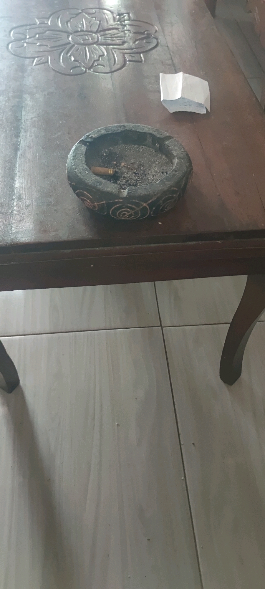 The image shows a close-up of a dark wooden table with an ashtray containing cigarette butts and ash. There is also a small, folded piece of white paper on the table. The table is resting on a light-colored tiled floor. The room appears to be indoors, possibly a common area in a home or establishment. The lighting is dim, casting shadows on the floor. The image does not contain enough information to determine the specific location within Gambarsari, Indonesia.