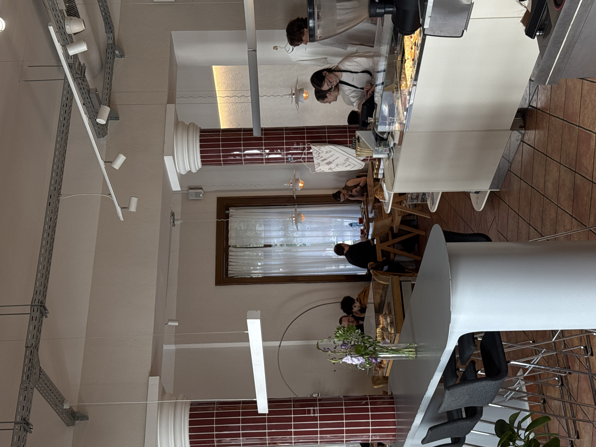 The image depicts the interior of a modern cafe, likely located in Belgrade, Serbia, with a clean and contemporary aesthetic. In the foreground, a long, white counter with high stools suggests a seating area for patrons. Behind the counter, staff members are visible, with one individual in a white shirt appearing to be a cashier or server. Behind them, a display case filled with pastries is evident.
Further into the cafe, several patrons are seated at tables, engaged in conversation or enjoying their meals. A large window with white curtains lets in natural light. The cafe's architecture features red-tiled cylindrical columns, adding a touch of color and contrast to the otherwise neutral palette. Exposed metal conduits are visible on the ceiling, contributing to the industrial-chic design. The overall atmosphere appears relaxed and social.