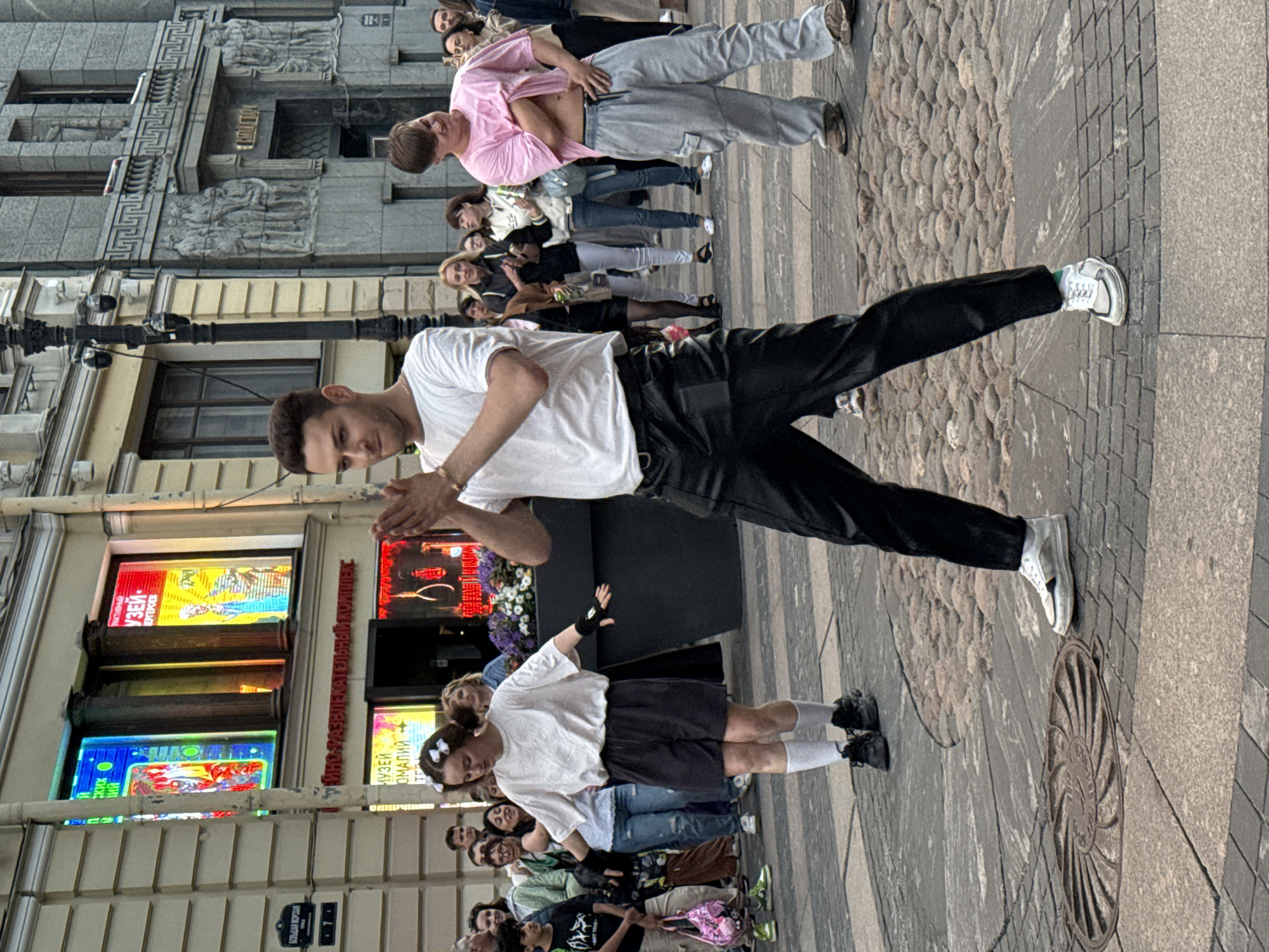 The image depicts a street performance in Saint Petersburg, Russia, likely on Nevsky Prospekt, indicated by the architectural style of the buildings and the prominent signage with Cyrillic text. In the foreground, a man with short brown hair, wearing a white t-shirt and black trousers, is captured mid-dance with his hands clasped. To his left, a woman with her hair in two buns, adorned with white bows, is also dancing, dressed in a white t-shirt, dark shorts, and white socks. To the right, a young man in a pink t-shirt and light grey cargo pants is performing, pulling up his shirt. A crowd of onlookers, dressed in casual attire, observes the performance. The street is paved with cobblestones and granite slabs, and a manhole cover is visible in the lower left corner. The background features a building with large windows displaying colorful imagery and Cyrillic signage, consistent with a museum or entertainment complex. The architecture suggests a historical district within the city.