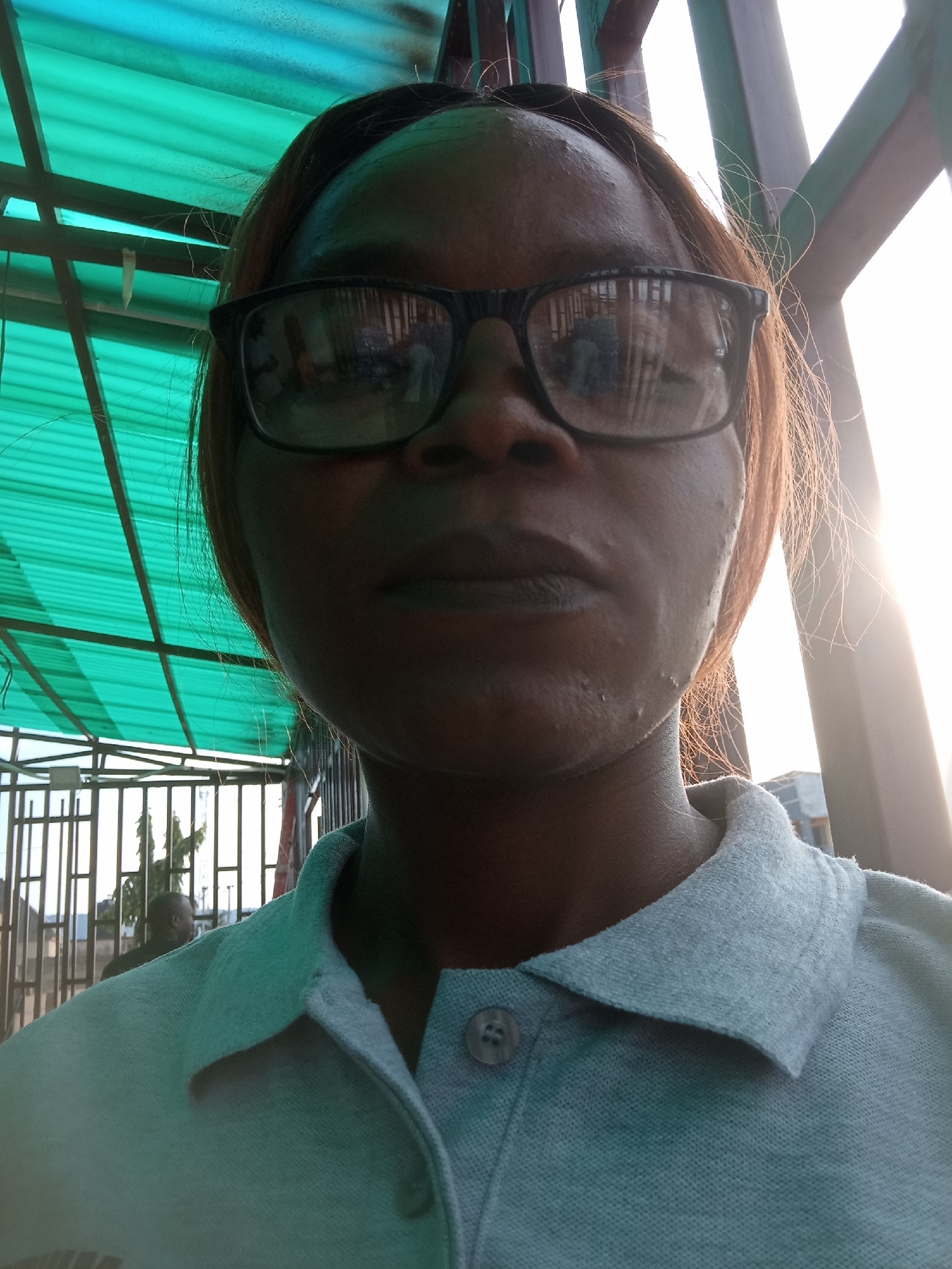 The image is a close-up selfie taken by a person wearing glasses and a light blue polo shirt. The person's face is the primary focus, with their head slightly tilted. Behind them, a green corrugated roof structure is visible, along with a metal fence and a bright, hazy sky. The lighting suggests it is daytime, with the sun setting or rising, casting a warm glow. There is a blurry figure of another person in the background, near the fence. The environment appears to be an outdoor, possibly urban setting, consistent with a location in Howayi, Nigeria, perhaps near a market or a covered walkway.