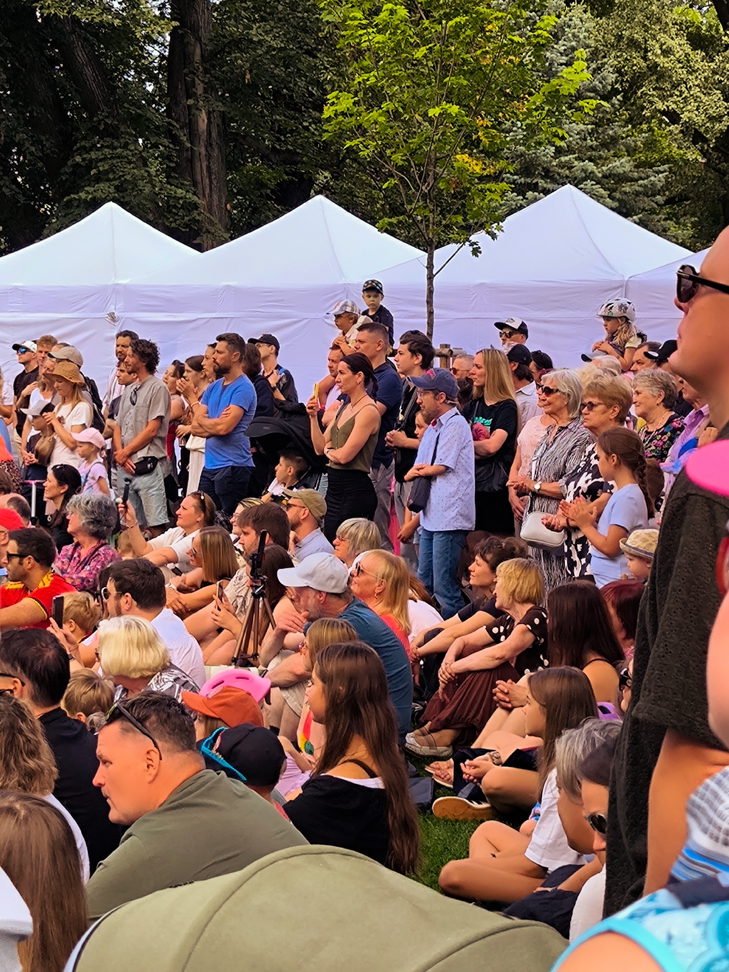 In Riga, Latvia, an outdoor event is underway, likely a festival or market, indicated by the white tents and a large gathering of people. The scene is populated by a diverse crowd, predominantly adults and some children, seated and standing on a grassy area. Many are dressed casually for warm weather.
In the foreground, a man in a green t-shirt is partially visible, looking to his left. To his right, several individuals are seated on the grass, including a woman in a white t-shirt and a younger girl with brown hair wearing a blue top. Further to the right, a woman in a black dress with white polka dots is visible, as is another woman in a black top.
In the mid-ground, the crowd continues, with individuals in casual attire. Some are standing and appear to be interacting. A man in a blue t-shirt stands with his arms crossed. Several women are also visible, some holding phones.
In the background, more white tents are set up, suggesting vendor stalls or performance areas. Tall trees with green foliage form a natural backdrop, indicating a park or outdoor public space. The overall atmosphere appears relaxed and festive, with many people observing something happening beyond the frame.