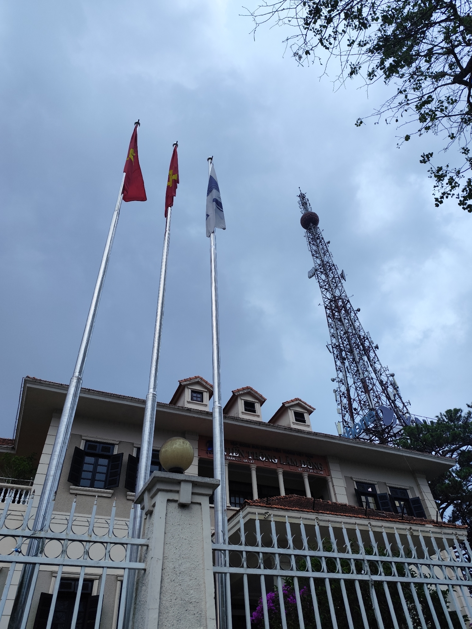 The image presents a low-angle view of a building, three flags, and a towering communications mast under an overcast sky. In the foreground, a white metal fence and a stone pillar with a spherical ornament partially obscure the lower facade of a cream-colored, multi-story building. This structure displays European-influenced architecture, featuring dark-shuttered windows, a tiled roof with dormer windows, and a second-story balcony.
Three tall, metallic flagpoles stand prominently in front of the building. Two display the red flag with a yellow star, identifying them as flags of Vietnam. The third flagpole on the right flies a white flag with a blue, stylized logo, possibly representing an organization.
Dominating the right side of the background is a massive lattice communications tower, densely packed with antennas and topped with a large, reddish-brown spherical component. Dark tree branches with some foliage frame the upper right corner of the sky.
Visible text on the building's facade reads "VIEN THONG LAM DONG" and below that "TRUNG TAM DA LAT." This translates to "Lam Dong Telecommunications" and "Da Lat Center," indicating the building likely houses a telecommunications facility. The cloudy, gray sky suggests an overcast day. The presence of the Vietnamese flags and the text confirms the location in Vietnam, specifically Da Lat, Lam Dong province. No people are visible, and no specific activity is taking place.