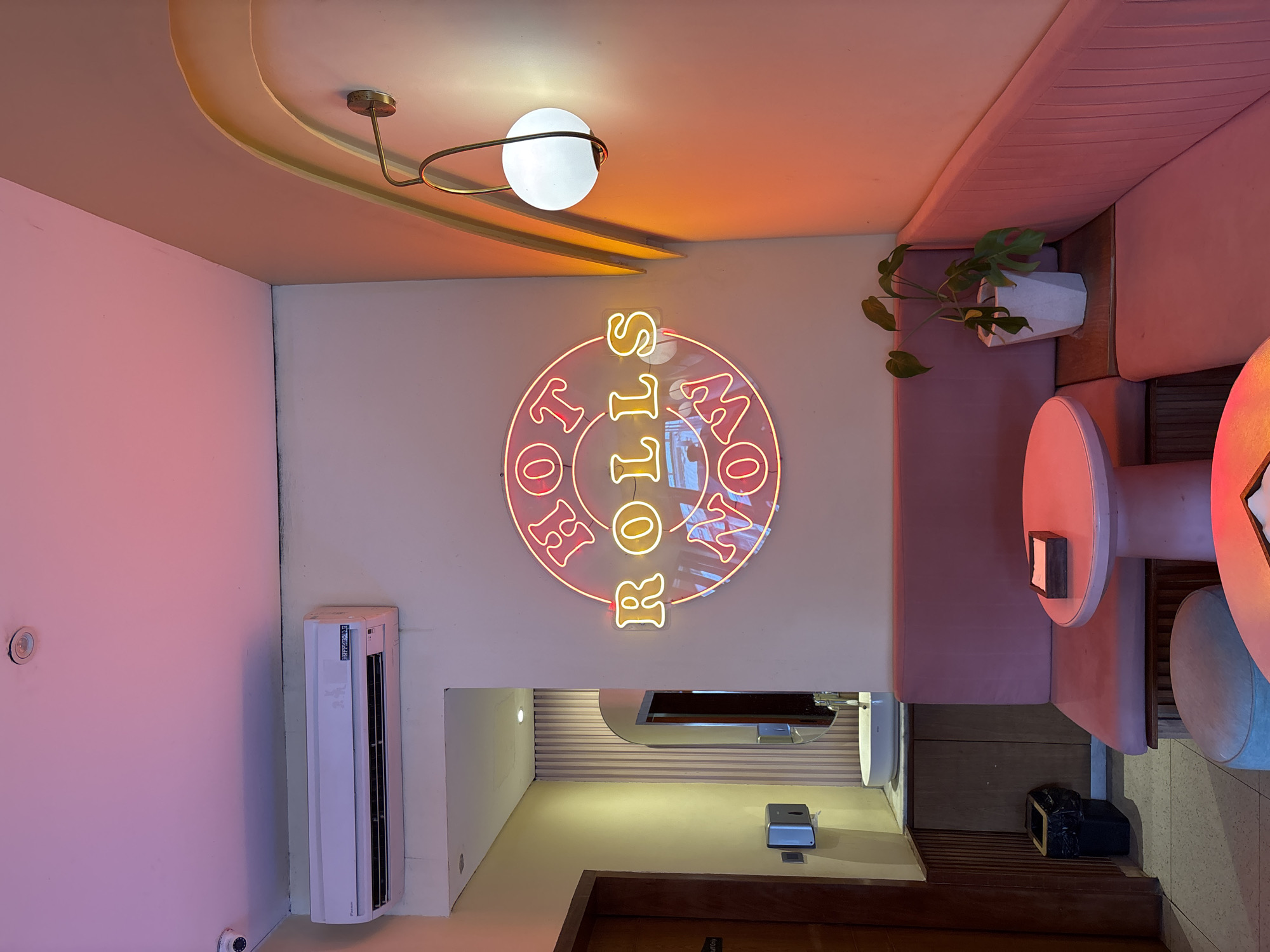 The image depicts the interior of a cafe or restaurant in Kuta Selatan, Indonesia, bathed in warm, ambient lighting, primarily from pink and orange hues. A prominent neon sign on the wall reads "HOT ROLLS NOW" in a circular arrangement with "HOT" and "NOW" in pink, and "ROLLS" in yellow. The seating area features plush pink banquette seating along the walls, with round pink tables and stools. A potted plant with green leaves sits on a wooden shelf between the banquettes. In the background, to the left, is a glimpse of a restroom area with a sink, mirror, and a hand soap dispenser. An air conditioning unit is mounted high on the wall above this area. A security camera is visible on the ceiling in the upper left corner. The overall aesthetic is modern and inviting, with a focus on creating a cozy and vibrant atmosphere.