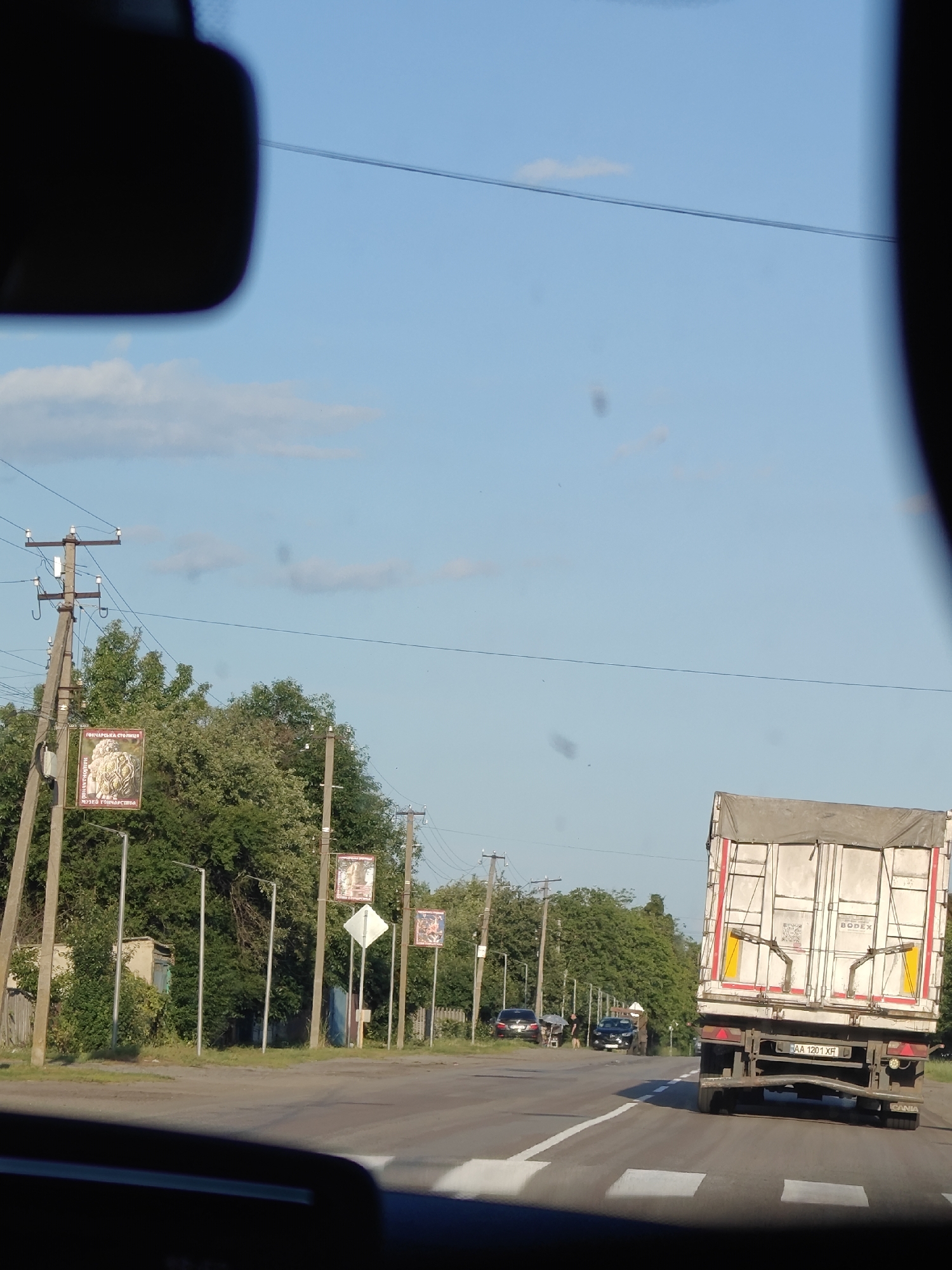 This media file captures a view from a vehicle, looking out at a roadside scene in Opishnia, Ukraine. The time of day appears to be late afternoon or early evening, as indicated by the warm, soft lighting and the clear blue sky with scattered clouds. The weather seems pleasant, suitable for outdoor activities.
The foreground is dominated by the interior of the vehicle, with a blurred rearview mirror and dashboard visible. The main subject of the image is the road ahead, with a white lane marker indicating the direction of travel. On the right side of the road, a large, white semi-trailer truck with red and yellow accents is prominently featured, moving away from the viewer. The truck has "BODEX" written on its side and a Ukrainian license plate reading "AA 1201 XF".
To the left of the road, a line of utility poles with electrical wires stretches into the distance. Several of these poles are adorned with rectangular posters or banners, some featuring images and text. One visible banner appears to be advertising the "MUSEUM OF POTTERY" in Opishnia, with text in Ukrainian. Further down the road, more poles with similar banners and streetlights are visible.
Behind the utility poles, lush green trees and dense foliage line the side of the road, suggesting a semi-rural or suburban setting. A few smaller buildings and a wooden fence are partially visible amongst the trees. In the distance, other vehicles are seen on the road, including cars and potentially other trucks. Two figures are standing near parked cars on the left side of the road, though their activity is unclear.
The overall impression is one of a typical day in a Ukrainian town, with everyday traffic and local advertisements on display. The scene is peaceful and ordinary, without any dramatic events or strong emotional cues.