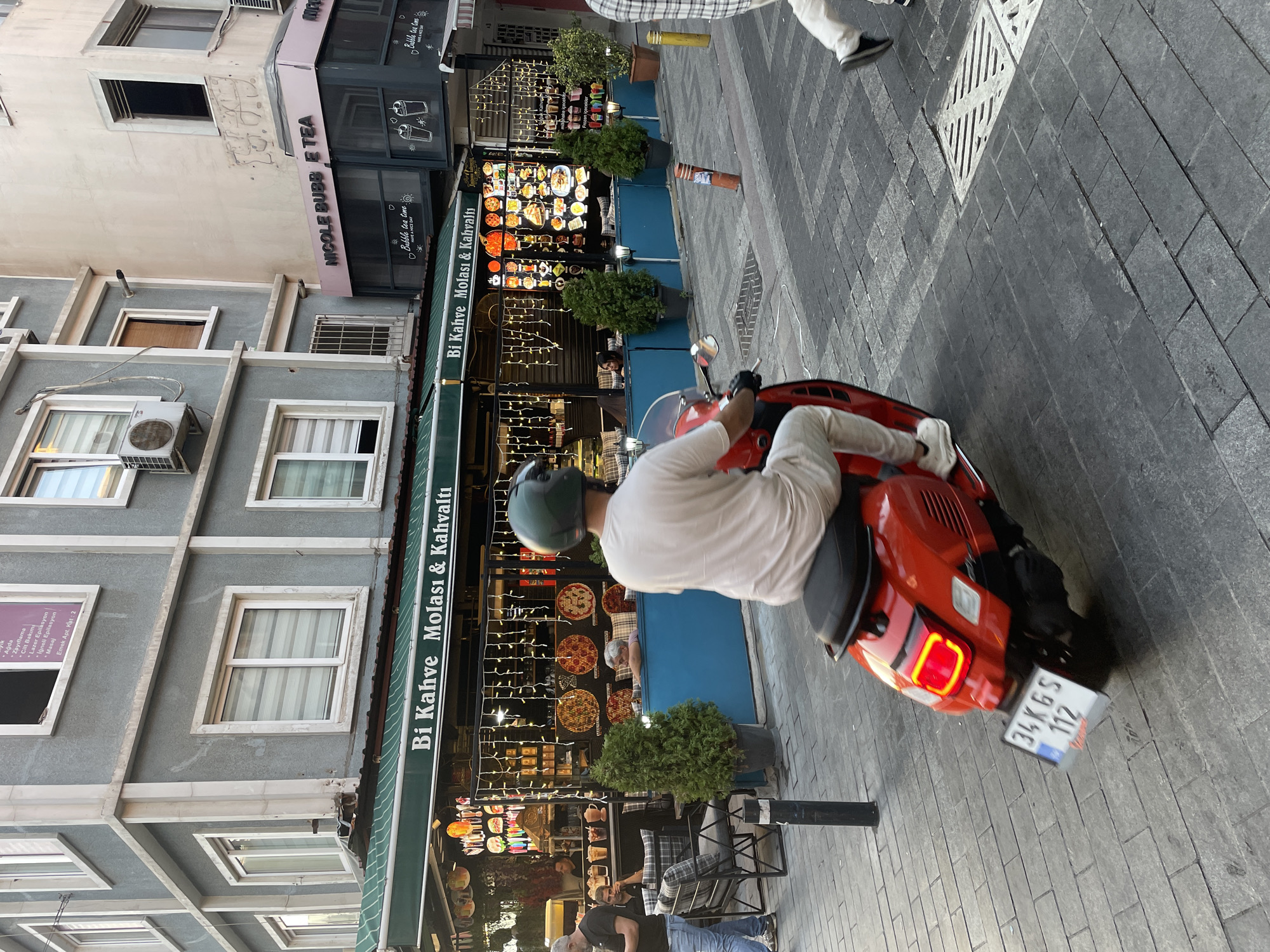 A red scooter with a license plate that reads "34 KGS 112" is being ridden by a person wearing a light-colored t-shirt and white pants, and a green helmet, on a cobblestone street in Kadıköy, Turkey. The scooter is in motion, and the rider's legs are visible. In the background, there is a cafe with a green awning that says "Bi Kahve Molası & Kahvaltı," meaning "A Coffee Break & Breakfast." The cafe has outdoor seating with blue seating cushions and decorative lights. Next to the cafe, there is a shop called "NICOLE BUBBLE TEA." The buildings in the background are multistory with windows. Other people are visible in the cafe and walking on the street. The overall scene depicts a typical street view in a bustling urban environment.