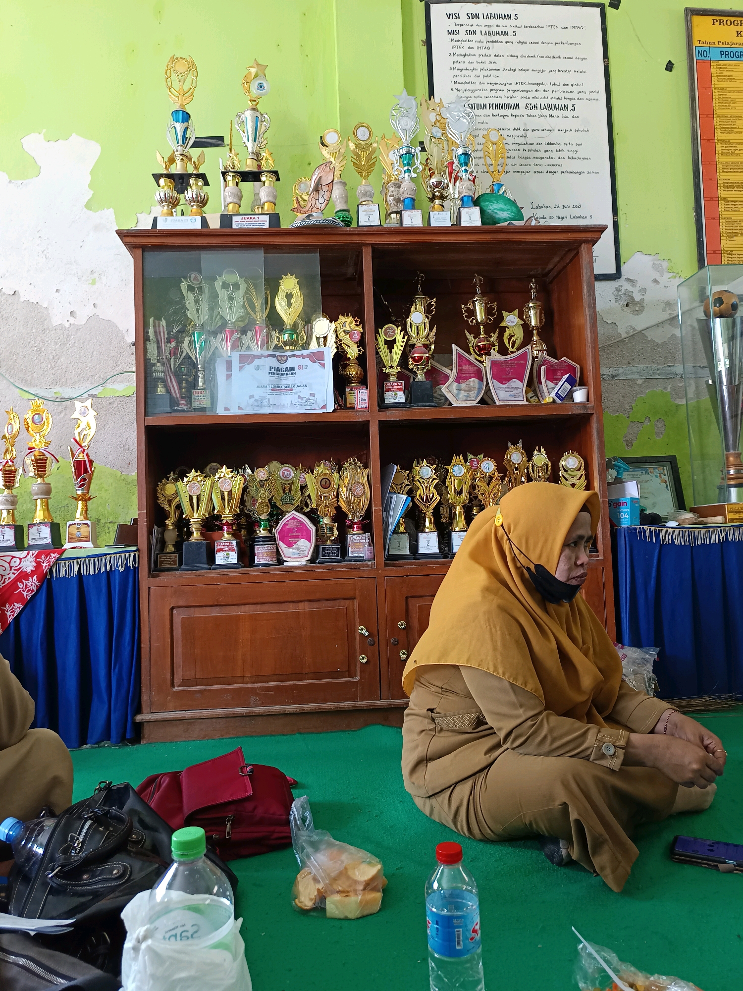The image depicts an indoor scene likely within a school in Labuhan, Indonesia. Dominating the background is a wooden cabinet filled with numerous trophies of varying sizes and designs, suggesting an award ceremony or a display of academic achievements. The cabinet has glass doors, revealing the trophies organized on shelves. Above the cabinet, a portion of a wall is visible, painted a light green with some peeling or chipped paint. A whiteboard with text, likely school-related information, is affixed to the wall. In the foreground, a woman wearing a tan uniform and a yellow hijab is seated on the green carpeted floor. She is facing to the right of the frame, and her body is partially obscured by objects in the foreground. Beside her, on the floor, are several items including a red bag, a black handbag, two plastic water bottles, and a plastic bag containing what appear to be snacks. The lighting suggests an indoor setting, possibly during daytime.