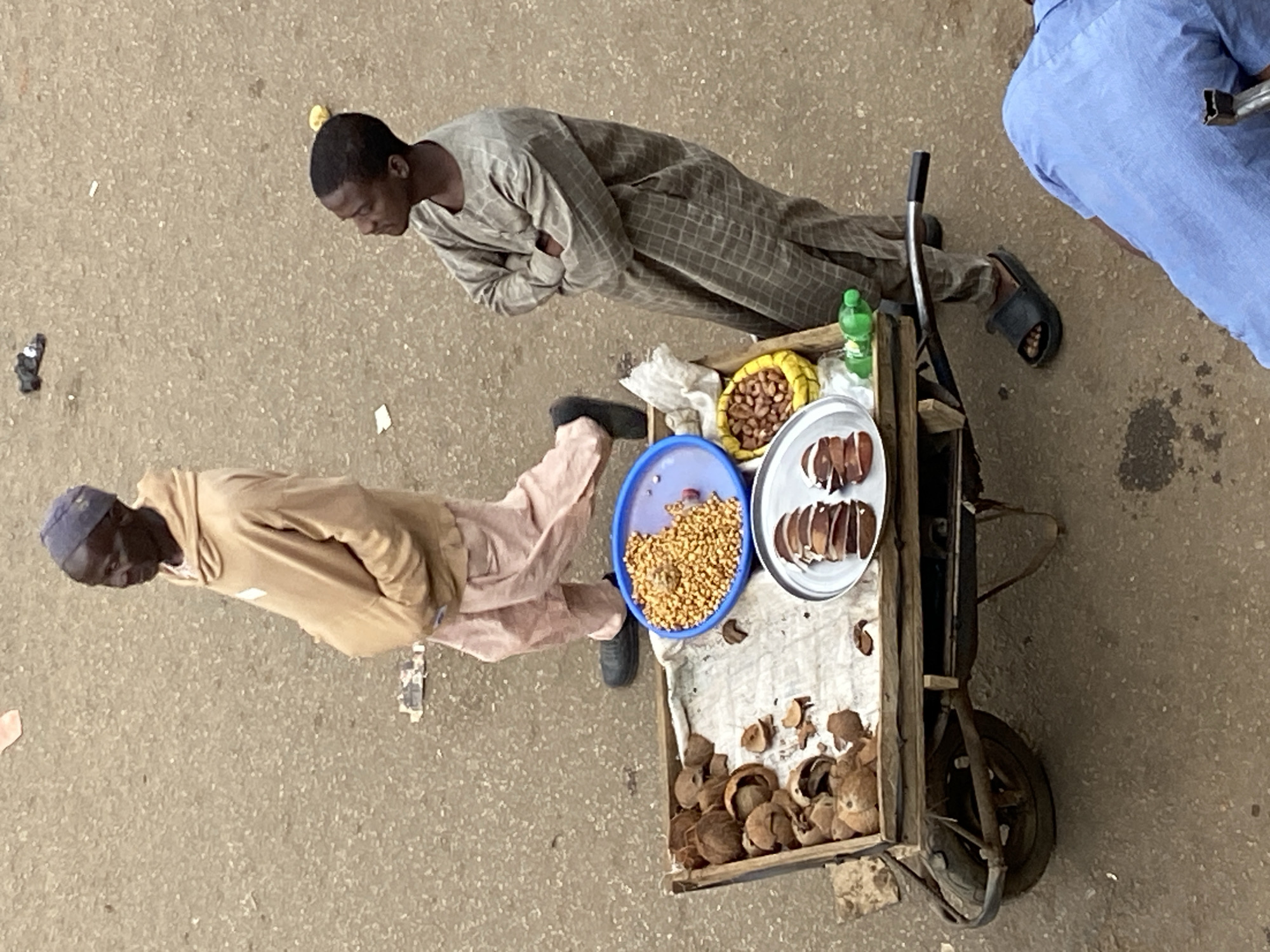 An overhead shot captures a street vending scene on a paved surface, likely in Bukuru, Nigeria, during the daytime. Two men are prominently featured alongside a wooden handcart laden with food items.
One man, positioned on the left, wears a light grey, grid-patterned traditional robe with long sleeves, and black sandals. He stands with his arms crossed, looking downwards towards the cart. Closer to the cart stands another man, wearing a tan hoodie, light-colored trousers, and a blue cap. He is also looking down, and his black shoe is visible near the cart. A third person, wearing a blue shirt, is partially visible on the far right of the frame.
The handcart, featuring a rustic wheel on the right, serves as a display for the vendor's goods. On its surface, a blue bowl contains a generous pile of small, light brown, granular items, resembling roasted peanuts or a similar snack. Adjacent to it, a yellow woven basket holds larger, yellowish-brown nuts. A silver-colored plate displays several oval, sliced segments, possibly nuts or fruit. A small green plastic bottle stands near these items. The front part of the cart's display area is covered by a piece of light-colored fabric and is littered with numerous discarded shells, suggesting they are the remnants of the products being sold, such as kola nuts or other local snacks.
The ground is a speckled light brown, paved surface, indicating an outdoor setting, possibly a street or market pathway. The bright, even lighting suggests it is mid-day. No visible text is present in the image.