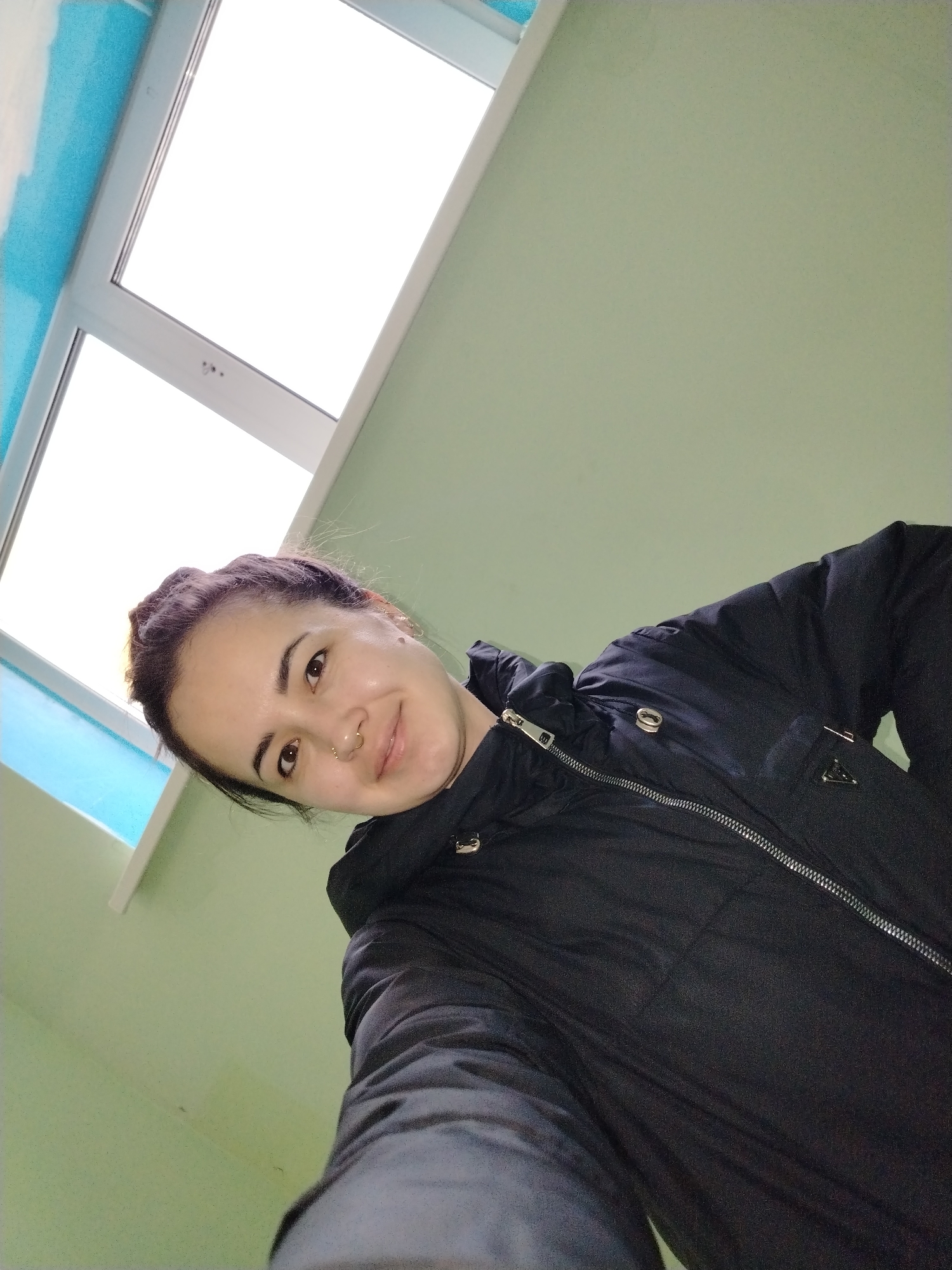 A woman is taking a selfie in what appears to be an interior space. She is wearing a black jacket with a zipper. The background consists of a light green wall and a window frame, with the window showing an overcast sky. The location is Noviy, Russia.