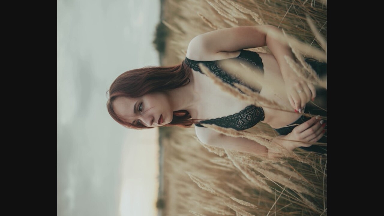 A young woman with auburn hair stands in a field of dry, tall grass, looking to her right. She is wearing a black lace bralette and dark bottoms. The sky is overcast and a soft light illuminates the scene. The grass is golden and the wind appears to be blowing through it.