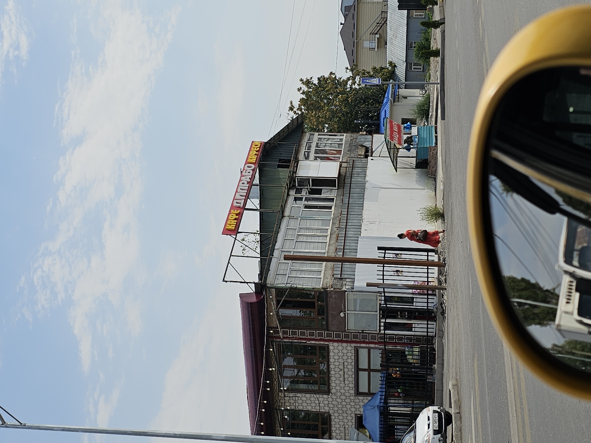 A street-level, eye-level shot captures a commercial building in the city of Özgön, Kyrgyzstan, on a clear day with a few scattered clouds. The building, seemingly a cafe, is two stories high and features a red awning with yellow Cyrillic lettering that reads "КАФЕ ЛИГРАБО КАФЕЧИ". The facade is a mix of textured stone or tile and modern windows, with some areas appearing to be under construction or renovation, evidenced by exposed metal framing and corrugated metal siding. A black metal fence runs along the front of the property, with a blue tent and some decorative items visible behind it. To the right of the main building, a smaller kiosk with a blue roof and red signage stands. A woman in a red dress is walking away from the building and towards the right side of the frame, carrying a dark bag. A white car is parked on the left, and its license plate is partially visible. In the foreground, the yellow edge of a car's rearview mirror is prominently featured, reflecting the road, other vehicles, and trees. The street in front of the building is asphalt with painted white lane markings. Residential buildings can be seen in the background.