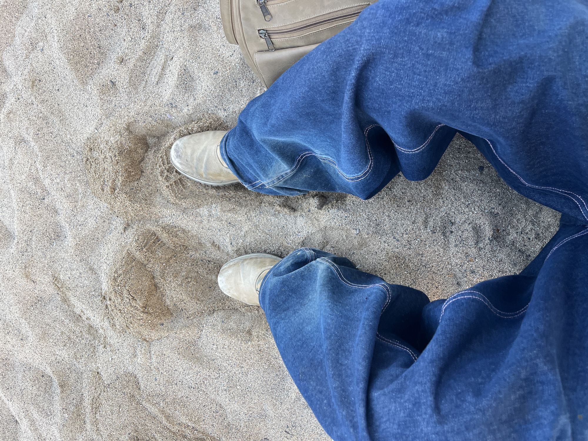 The image shows a first-person perspective of a person sitting on a sandy surface, likely a beach. The person is wearing loose-fitting dark blue jeans and light-colored slip-on shoes. A tan-colored bag with two zippers is visible to the right of the frame. The sand is textured with visible grains and some small pebbles. The lighting suggests it is daytime. There is no information in the image to indicate the location is the city of Barcelona, Spain.