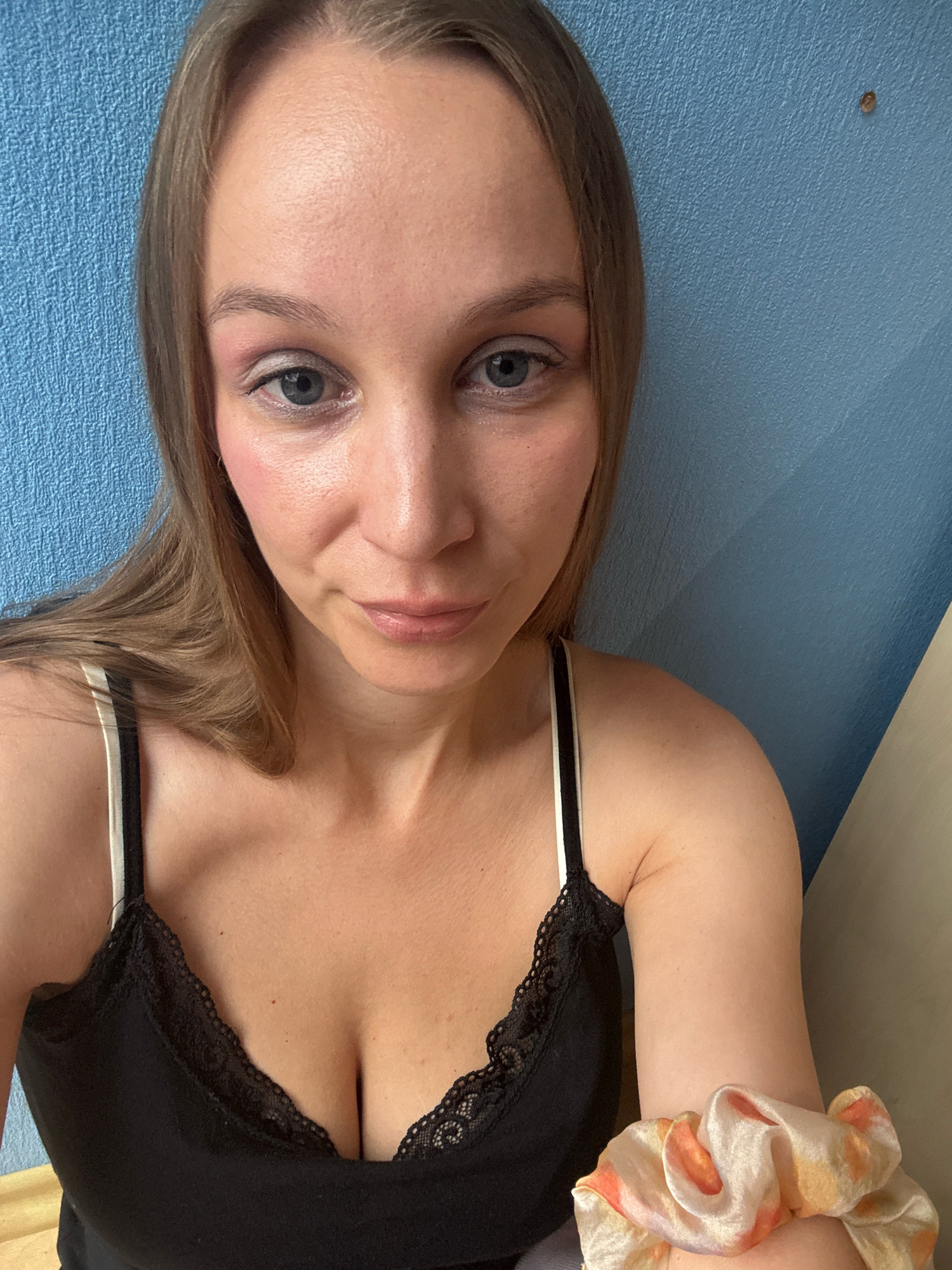 A close-up selfie shows a woman with light brown hair and blue eyes. She is wearing a black camisole with lace trim and a white and beige scrunchie on her right arm. The background is a textured blue wall. The image does not contain any information indicating the location in Überherrn, Germany.