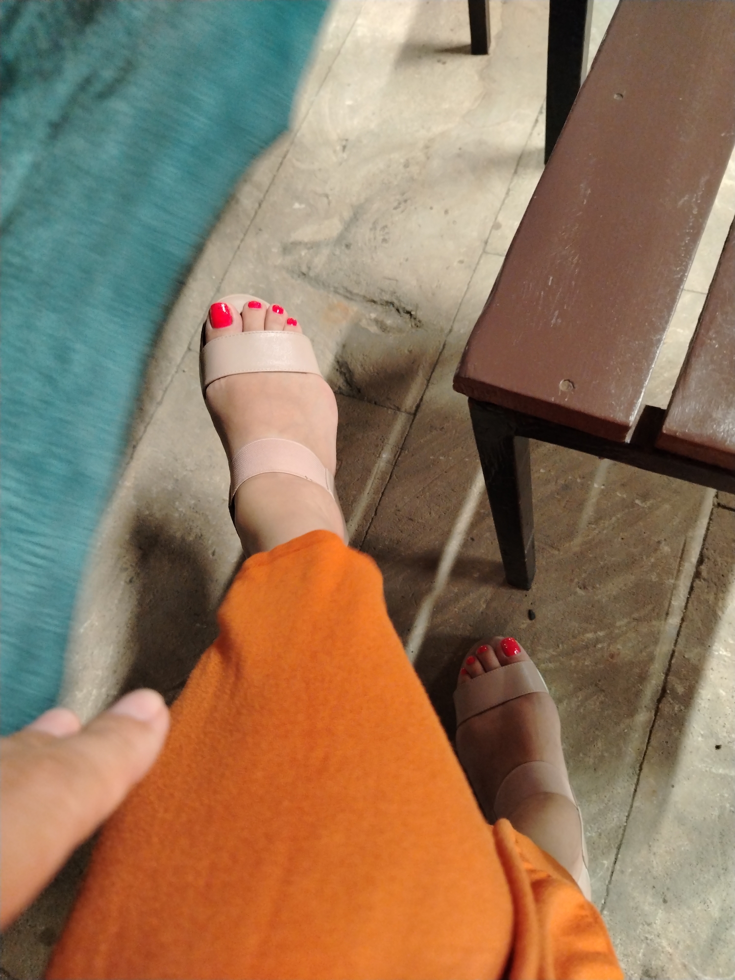 In an outdoor setting with a tiled floor, two feet are visible, both adorned with bright red nail polish. The left foot is partially covered by a folded orange garment, and a thumb is just visible in the lower left corner, appearing to be touching the fabric. This foot is wearing a beige sandal with two straps across the toes and one around the ankle. The right foot is also wearing a similar beige sandal. In the background, a brown wooden bench with dark legs is present, and to the left, a textured blue surface, possibly a pool or a water feature, is visible. The scene suggests a casual moment, potentially in a public area like a park or a poolside cafe. The lighting indicates it might be daytime, possibly late afternoon or evening due to the shadows. There is no specific information to suggest this location is in Sumqayıt, Azerbaijan.