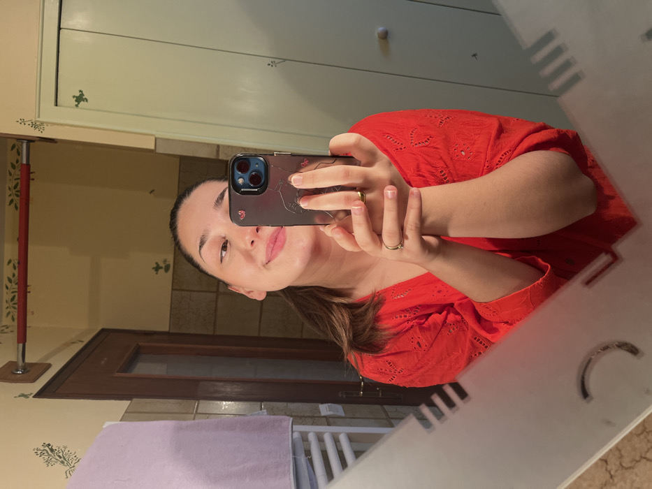 A female individual is depicted in a mirror selfie, centered from the upper chest upwards, within an indoor setting in Ancenis, France. She possesses dark hair pulled back and a smiling facial expression. She is wearing a red, long-sleeved top featuring eyelet embroidery. A gold-colored ring is visible on the ring finger of her left hand. She holds a dark-colored smartphone, equipped with a triple-lens camera array, in both hands, directing it towards the mirror for the photograph.
The reflected background reveals elements consistent with a bathroom or dressing area. To the left, a white heated towel rail is partially visible, draped with a light purple towel. Behind the subject, a dark brown wooden door with a translucent upper panel is present. Above the door, a red-accented horizontal bar is mounted on a light-colored wall. The walls are a light beige or cream shade, adorned with repetitive green stenciled botanical patterns. To the right, a light green cabinet or closet with two circular light-colored knobs is visible, also featuring similar green stenciled motifs. The mirror itself exhibits a frosted or etched geometric decorative pattern on its lower edges. The visible floor in the bottom left reflects as light-colored tiling.