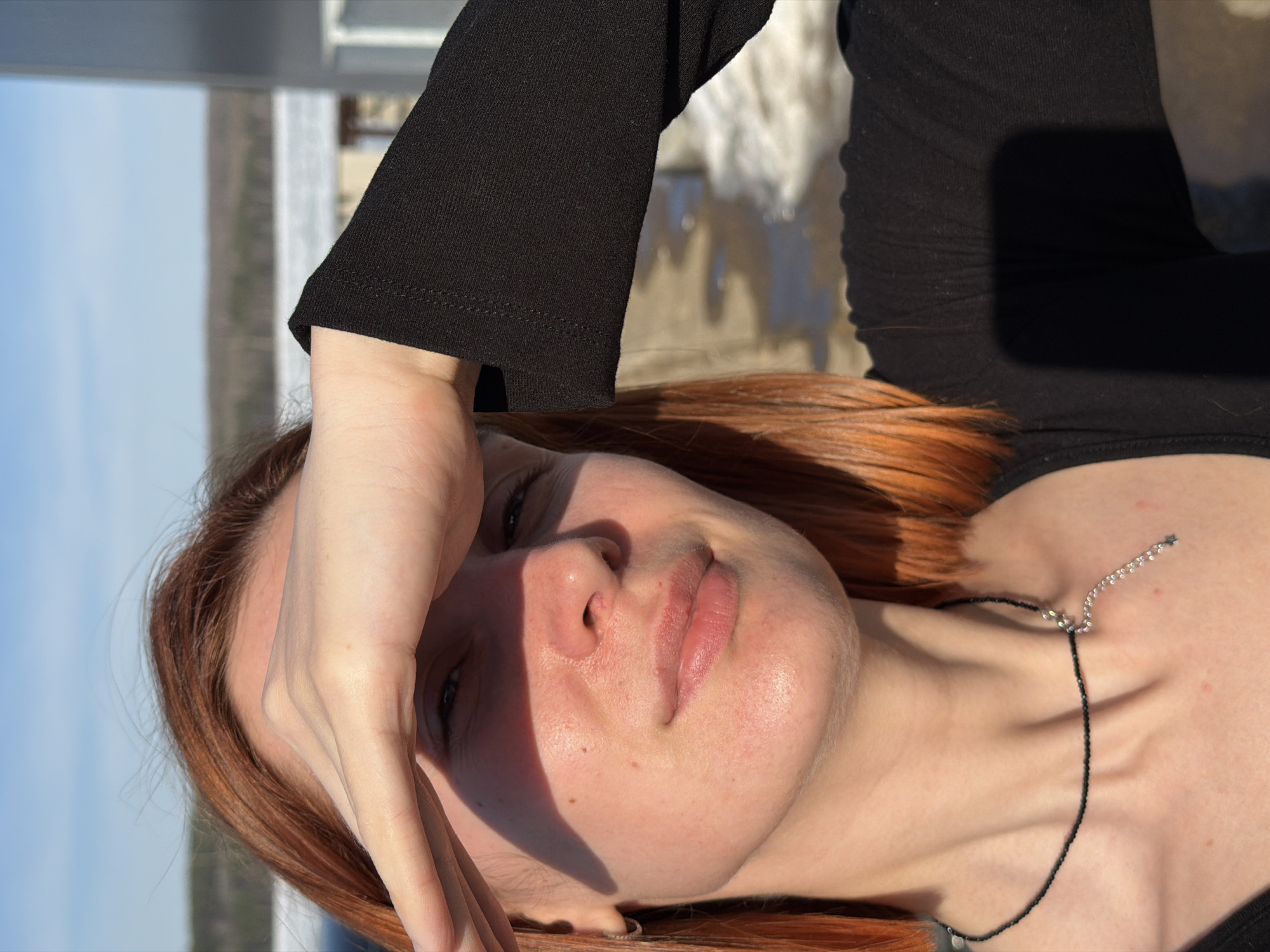 A woman with red hair is shielding her eyes from the sun with her hand. She is wearing a black top and a necklace. The background is a blurred landscape. This image may be taken in the city of Chaikovsky, Russia, although location cannot be confirmed with certainty from visual information.